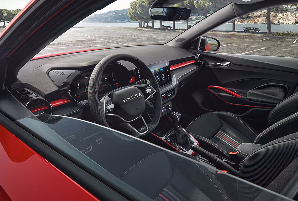 Wir blicken durch das geöffnete Fenster der Fahrertür in das Interieur des Škoda Fabia Monte Carlo in Velvet-Rot Metallic. Das Cockpit überzeugt mit schwarzen Sportsitzen im Streifendesign, einem Multifunktions-Sportlenkrad und dynamischen roten Akzenten an Armaturentafel und Mittelkonsole. Durch die Frontscheibe ist eine sonnige Uferpromenade zu erkennen.