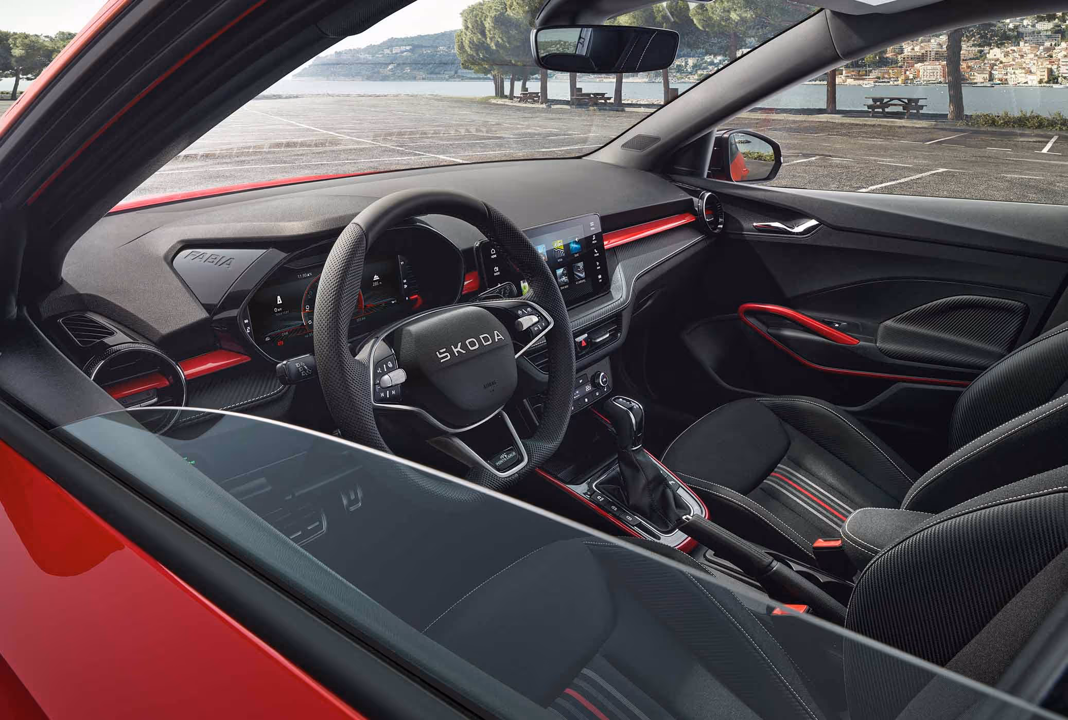 Wir blicken durch das geöffnete Fenster der Fahrertür in das Interieur des Škoda Fabia Monte Carlo in Velvet-Rot Metallic. Das Cockpit überzeugt mit schwarzen Sportsitzen im Streifendesign, einem Multifunktions-Sportlenkrad und dynamischen roten Akzenten an Armaturentafel und Mittelkonsole. Durch die Frontscheibe ist eine sonnige Uferpromenade zu erkennen.
