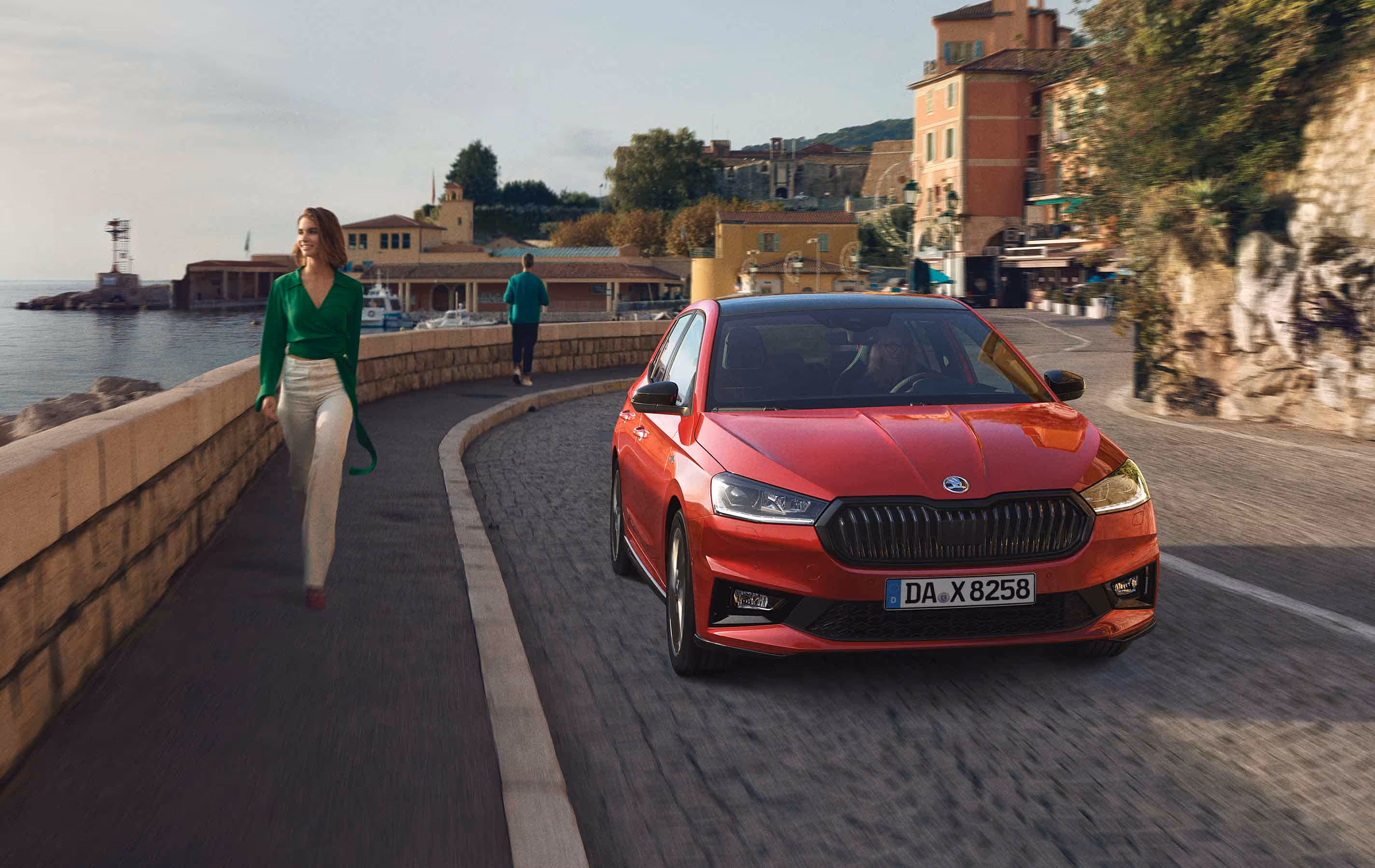 Ein Škoda Fabia Monte Carlo in der Lackierung Velvet-Rot Metallic fährt auf einer gepflasterten Küstenstraße, begleitet von einer Passantin auf dem angrenzenden Gehweg. Die sportliche Ausstattungslinie zeichnet sich durch markante schwarze Elemente wie den Kühlergrill und das Dach vor der mediterranen Kulisse aus.