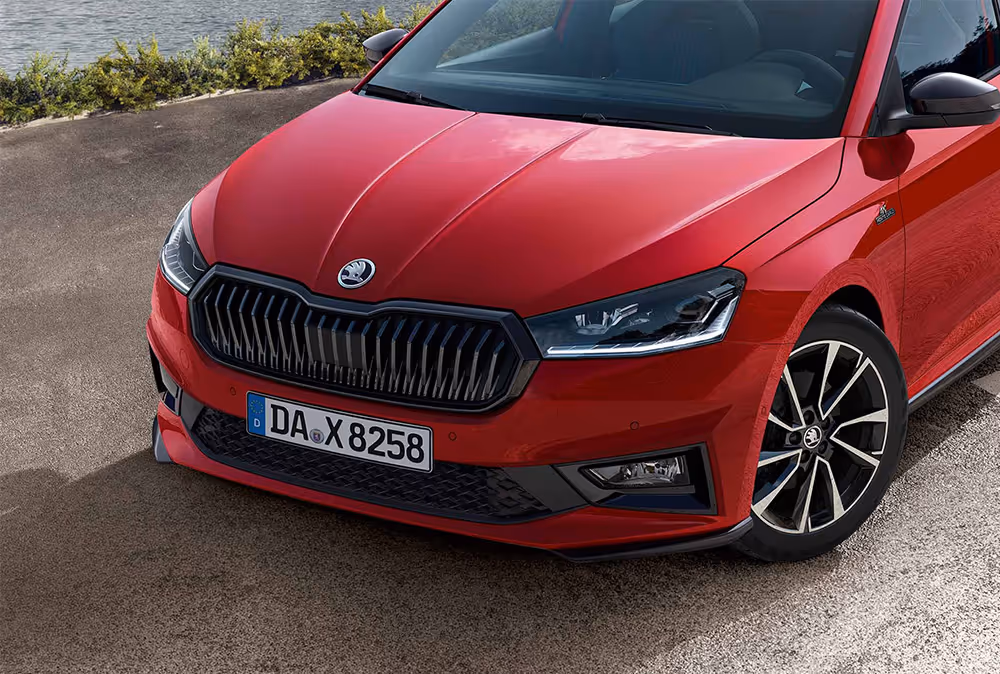 Eine Detailansicht der Frontpartie zeigt den Škoda Fabia Monte Carlo in der Lackierung Velvet-Rot Metallic vor einer Küstenkulisse. Markante Designelemente wie der schwarze Kühlergrillrahmen, die schwarzen Außenspiegelkappen und die Frontspoilerlippe unterstreichen den sportlichen Auftritt. Abgerundet wird das Profil durch die glanzgedrehten Leichtmetallfelgen und die Monte Carlo-Plakette am Kotflügel.