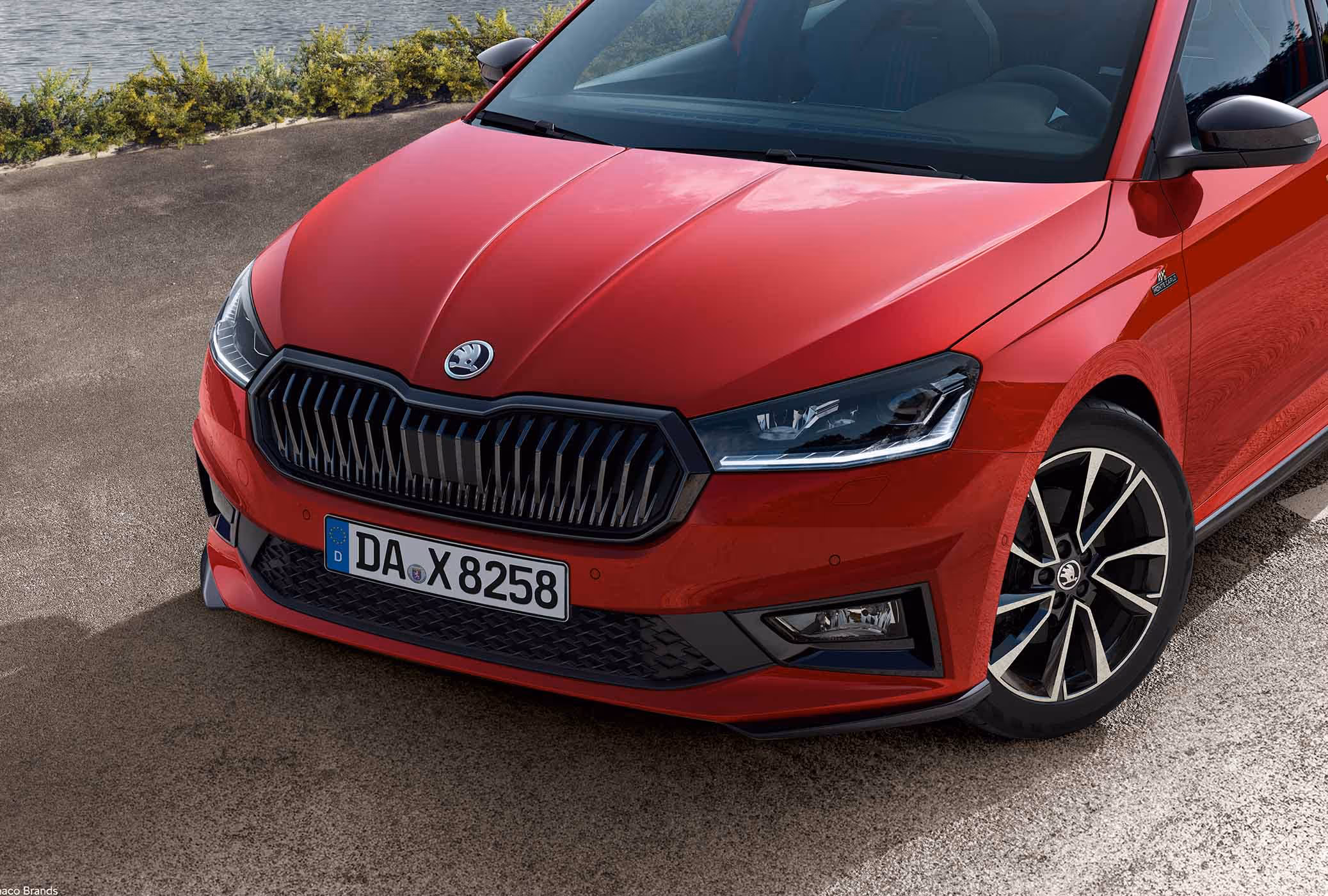 Eine Detailansicht der Frontpartie zeigt den Škoda Fabia Monte Carlo in der Lackierung Velvet-Rot Metallic vor einer Küstenkulisse. Markante Designelemente wie der schwarze Kühlergrillrahmen, die schwarzen Außenspiegelkappen und die Frontspoilerlippe unterstreichen den sportlichen Auftritt. Abgerundet wird das Profil durch die glanzgedrehten Leichtmetallfelgen und die Monte Carlo-Plakette am Kotflügel.

