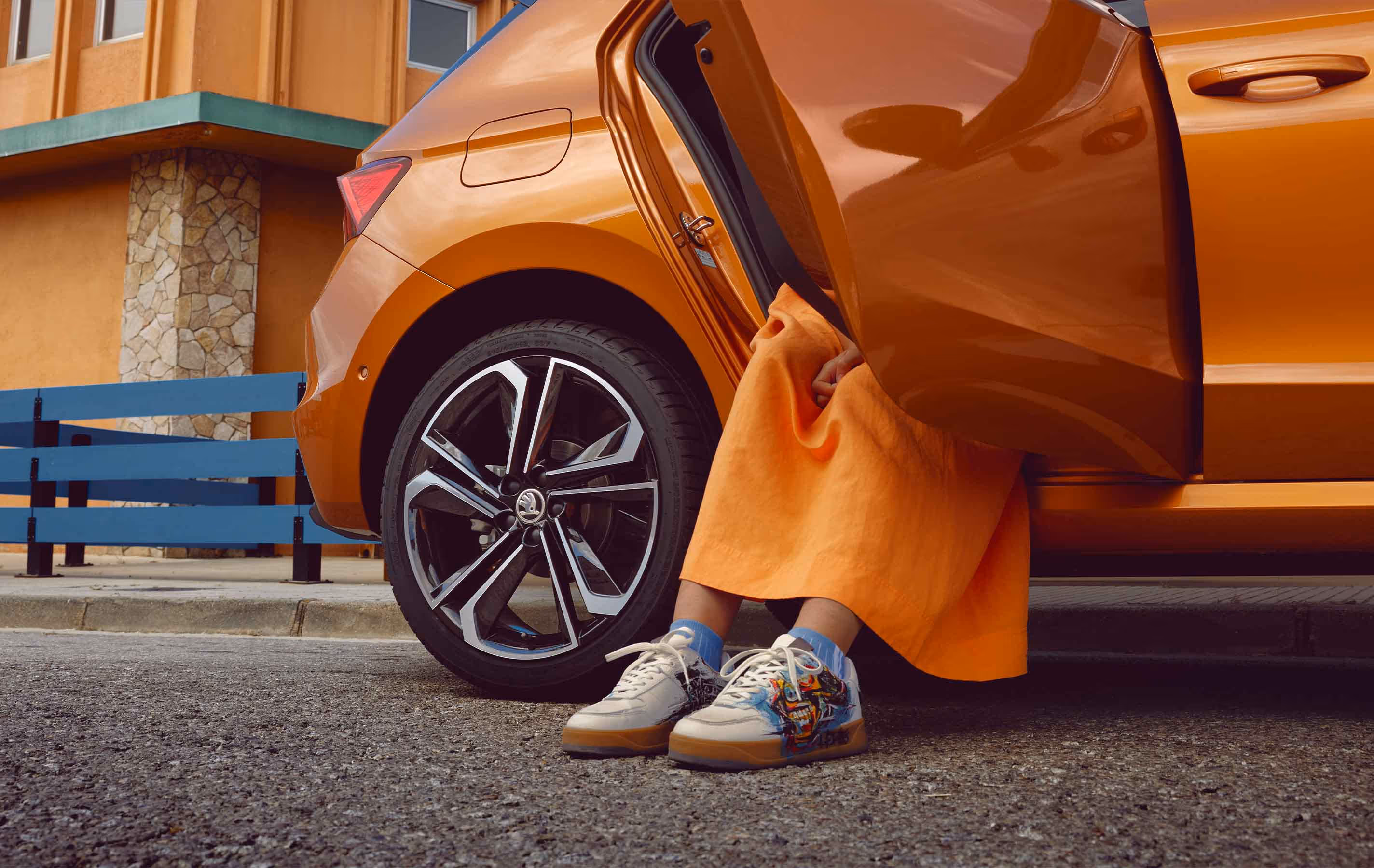Eine Detailaufnahme zeigt den hinteren Bereich eines Škoda Fabia in der Lackierung Phoenix-Orange Metallic, aus dessen geöffneter Fondtür eine Person in einem farblich abgestimmten Rock steigt. Im Fokus steht dabei die glanzgedrehte Leichtmetallfelge mit ihren markanten Aero-Einsätzen. Modische Sneaker und eine urbane Gebäudefassade im Hintergrund runden die Szenerie ab.