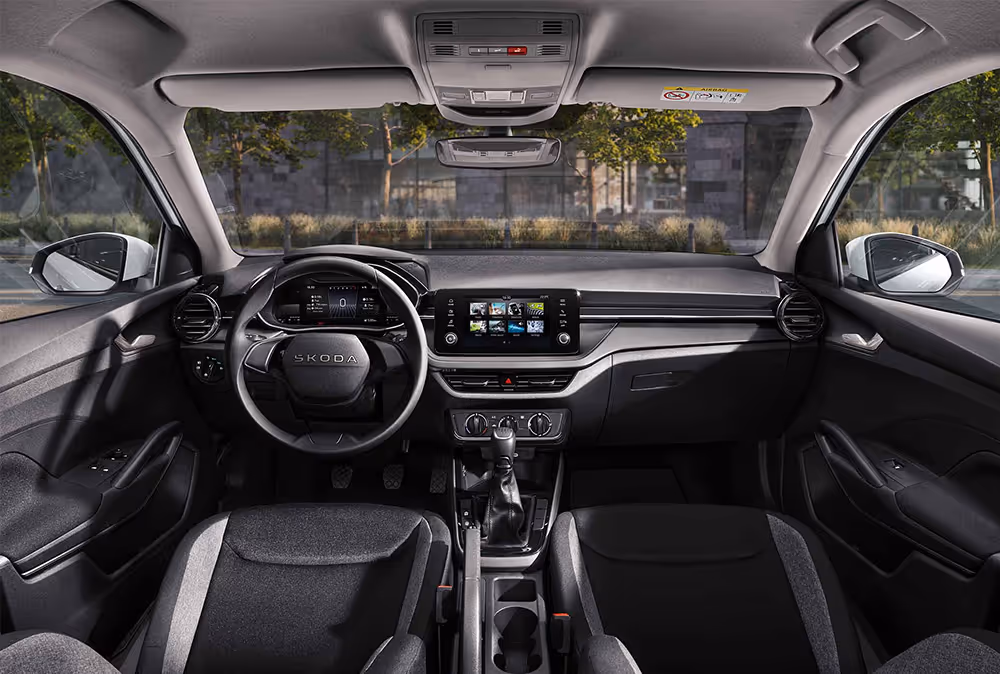 Eine weite Innenansicht zeigt das gesamte Cockpit eines Škoda Fabia in der Serienausstattung Essence. Das funktionale, dunkle Interieur ist mit schlichten schwarzen Stoffsitzen, einem griffigen Lenkrad und einem zentralen Infotainmentbildschirm ausgestattet. Das Armaturenbrett ist in genarbter Kunststoff-Optik gehalten. Durch die Windschutzscheibe ist eine städtische Kulisse mit Bäumen und einer Eisenbahnbrücke aus Naturstein zu sehen.