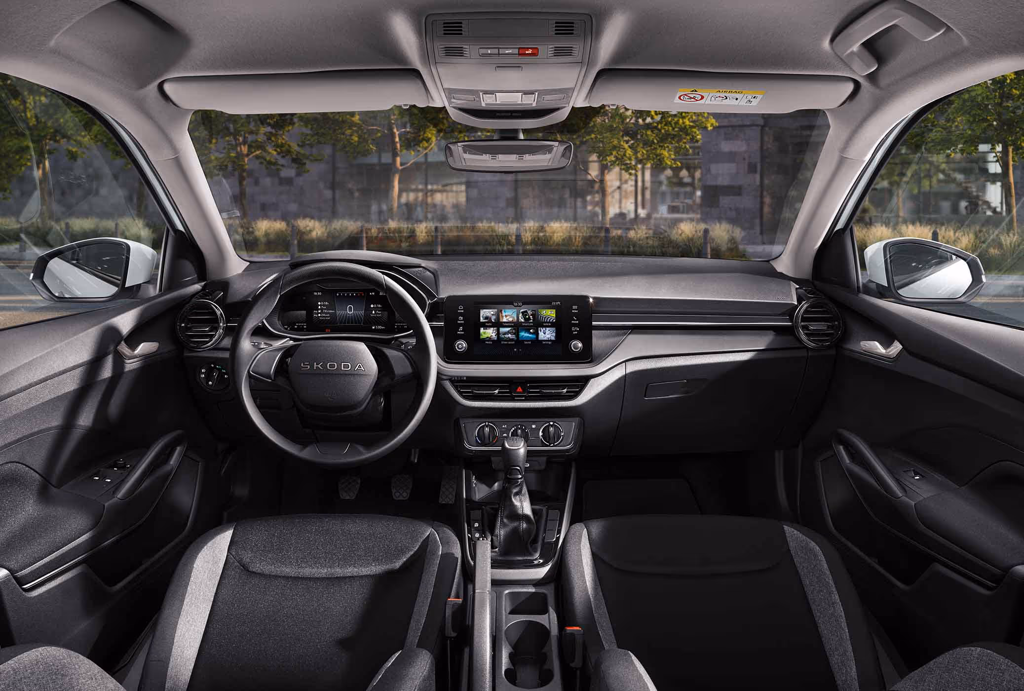 Eine weite Innenansicht zeigt das gesamte Cockpit eines Škoda Fabia in der Serienausstattung Essence. Das funktionale, dunkle Interieur ist mit schlichten schwarzen Stoffsitzen, einem griffigen Lenkrad und einem zentralen Infotainmentbildschirm ausgestattet. Das Armaturenbrett ist in genarbter Kunststoff-Optik gehalten. Durch die Windschutzscheibe ist eine städtische Kulisse mit Bäumen und einer Eisenbahnbrücke aus Naturstein zu sehen.