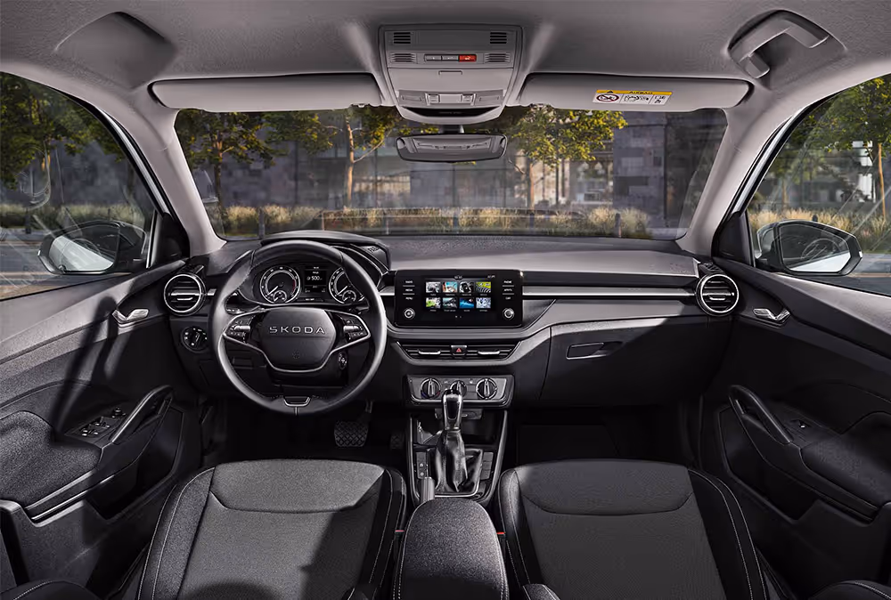 Eine weite Innenansicht zeigt das gesamte Cockpit eines Škoda Fabia mit der Design Selection Loft. Das puristische Interieur verfügt über schwarze Stoffsitze mit grauem Muster und Standard-Dekorelemente in Schwarz. Ein Infotainmentbildschirm und das Lenkrad stehen im Fokus des aufgeräumten Armaturenbretts. Durch die Windschutzscheibe ist eine städtische Kulisse mit Bäumen und einer Eisenbahnbrücke aus Naturstein zu sehen.