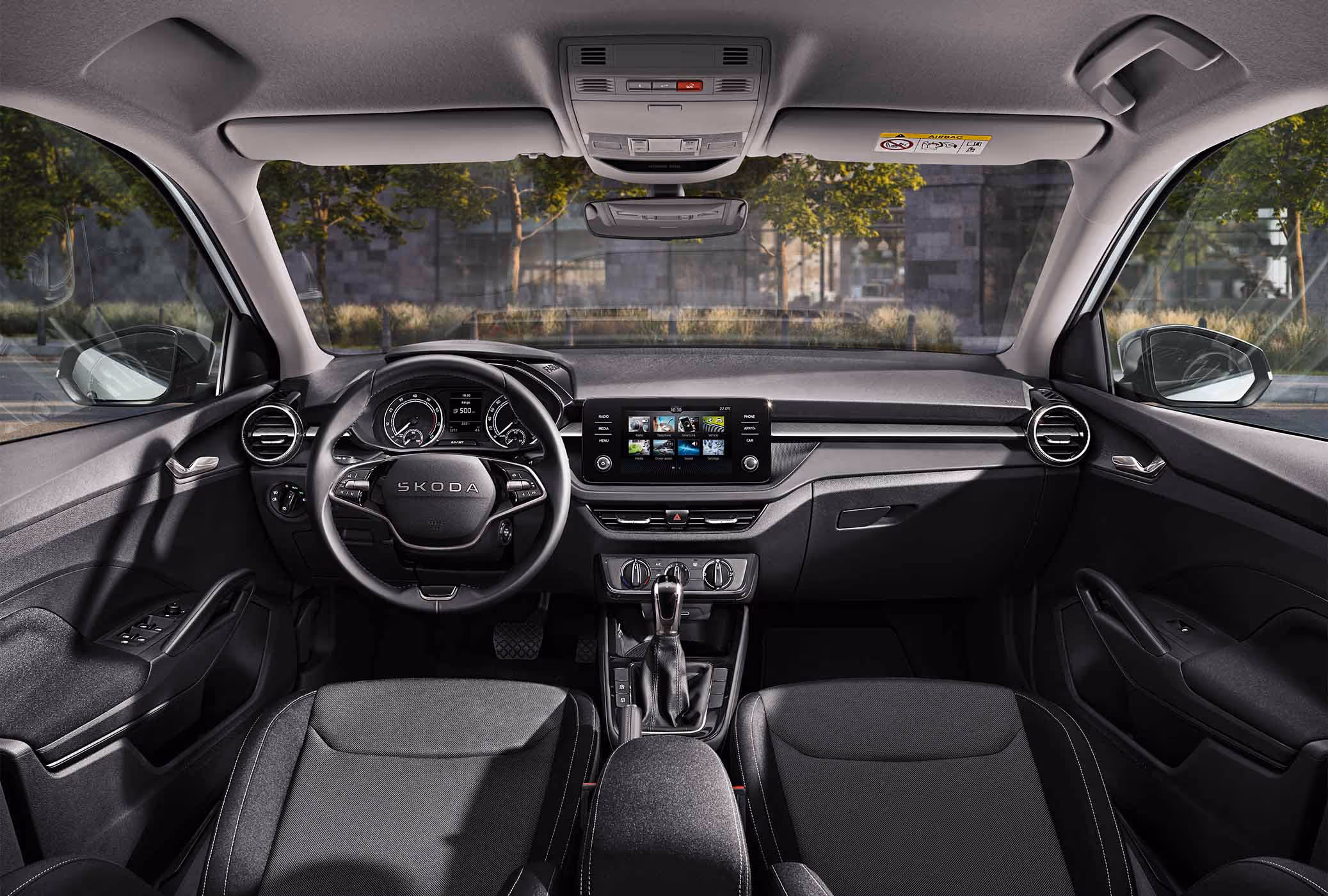 Eine weite Innenansicht zeigt das gesamte Cockpit eines Škoda Fabia mit der Design Selection Loft. Das puristische Interieur verfügt über schwarze Stoffsitze mit grauem Muster und Standard-Dekorelemente in Schwarz. Ein Infotainmentbildschirm und das Lenkrad stehen im Fokus des aufgeräumten Armaturenbretts. Durch die Windschutzscheibe ist eine städtische Kulisse mit Bäumen und einer Eisenbahnbrücke aus Naturstein zu sehen.