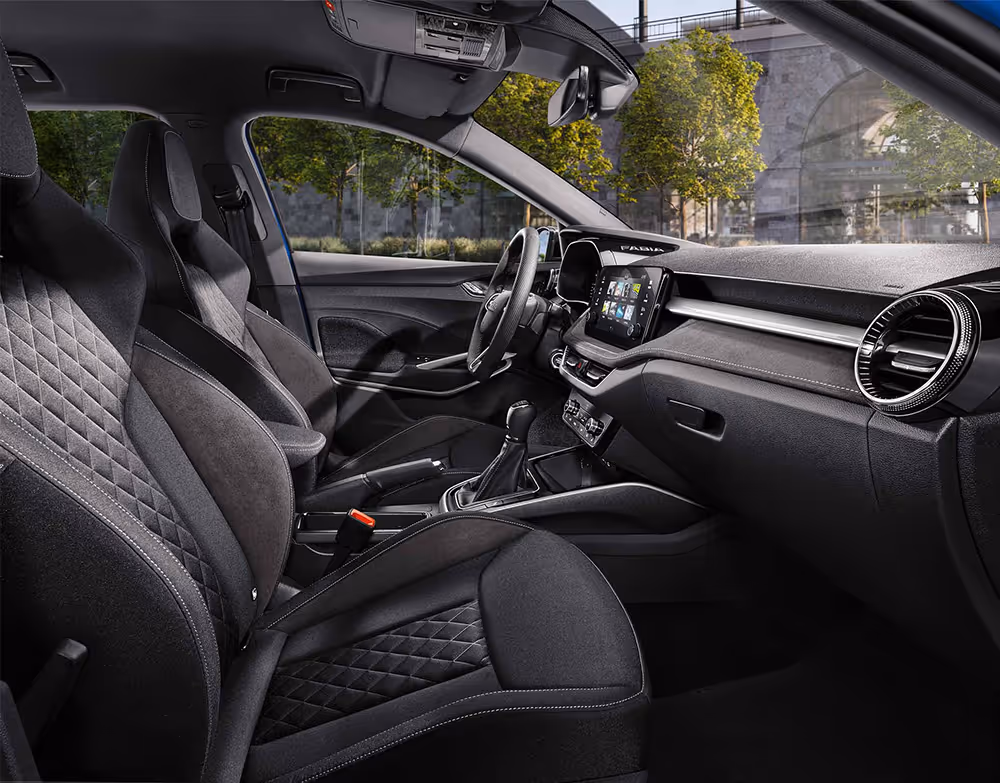 Eine seitliche Innenansicht des Škoda Fabia zeigt das Interieur der Design Selection Dynamic. Die schwarzen Sportsitze mit integrierten Kopfstützen und silbernen Nähten sowie der schwarze Dachhimmel sind zu sehen, während durch die Fenster eine städtische Kulisse mit Bäumen und einer Eisenbahnbrücke aus Naturstein sichtbar ist.