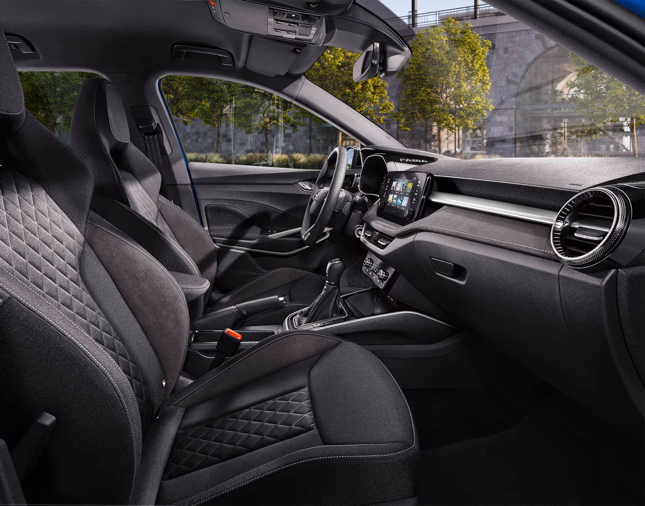 Eine seitliche Innenansicht des Škoda Fabia zeigt das Interieur der Design Selection Dynamic. Die schwarzen Sportsitze mit integrierten Kopfstützen und silbernen Nähten sowie der schwarze Dachhimmel sind zu sehen, während durch die Fenster eine städtische Kulisse mit Bäumen und einer Eisenbahnbrücke aus Naturstein sichtbar ist.