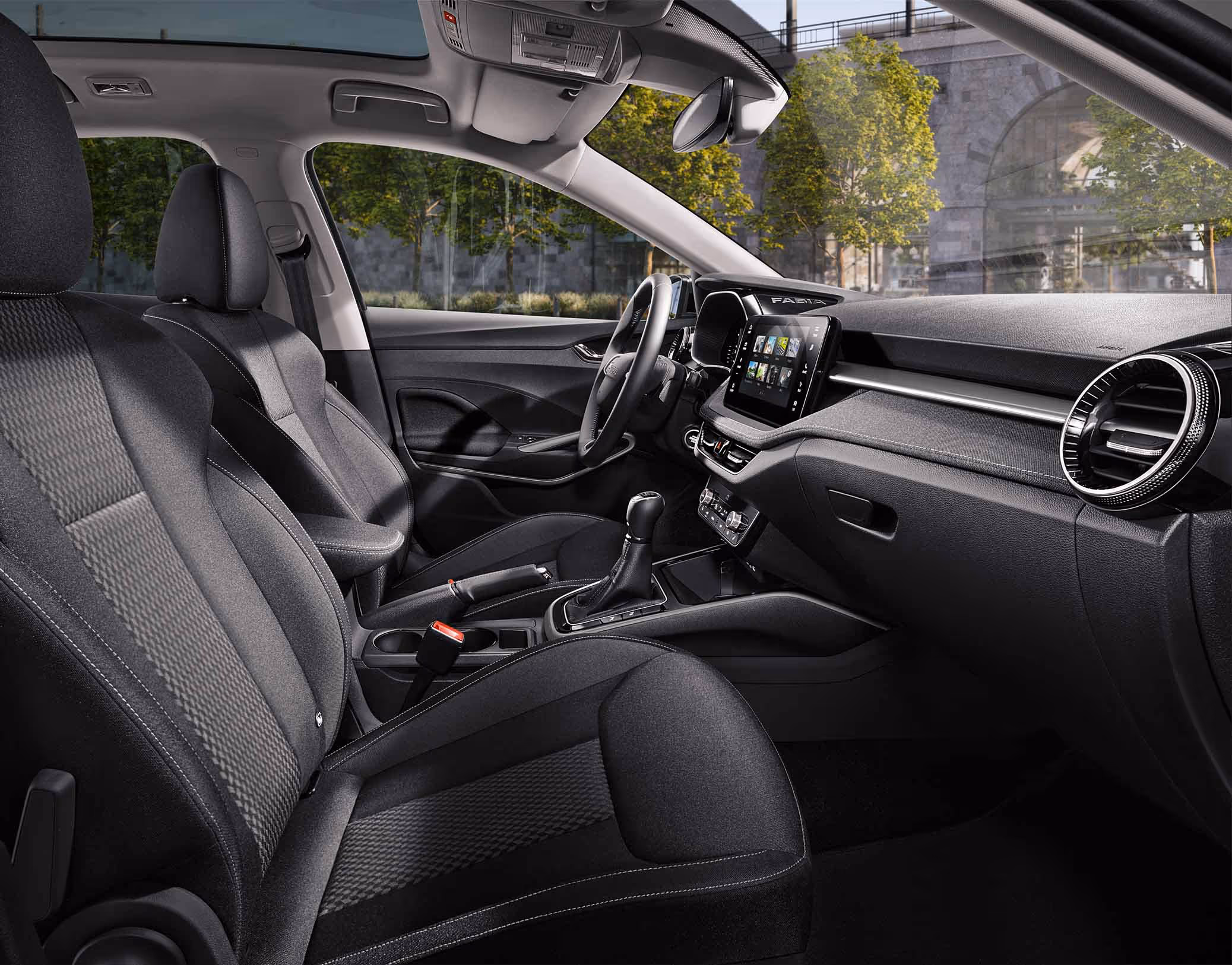 Eine seitliche Innenansicht des Škoda Fabia zeigt das Interieur der Design Selection Lodge. Die Sitzbezüge aus Suedia-Mikrofaser und Stoff mit kupferfarbenen Akzenten sowie das Armaturenbrett mit Dekor in Anodized Cross Copper sind zu sehen, während durch die Fenster eine städtische Kulisse mit Bäumen und einer Eisenbahnbrücke aus Naturstein sichtbar ist.