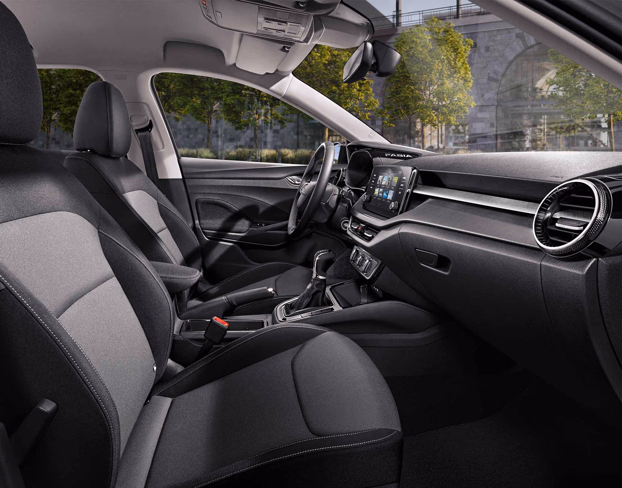 Eine seitliche Innenansicht des Škoda Fabia zeigt das Interieur der Design Selection Loft. Die grauen Stoffsitze mit grauen Kontrastnähten sowie die stoffbezogenen Elemente am Armaturenbrett sind zu sehen, während durch die Fenster eine städtische Kulisse mit Bäumen und einer Eisenbahnbrücke aus Naturstein sichtbar ist.