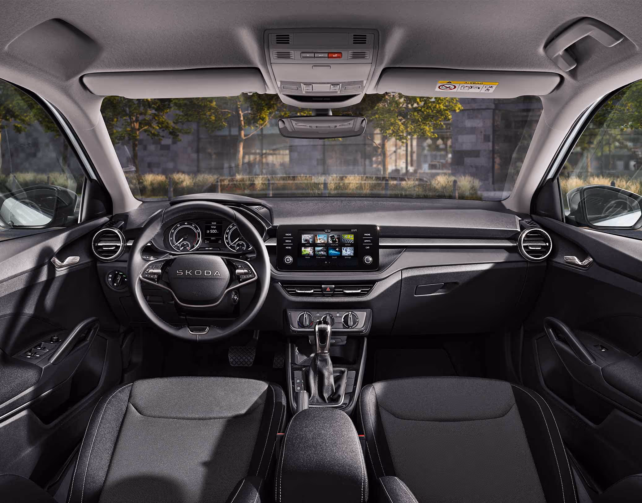 Eine weite Innenansicht zeigt das gesamte Cockpit eines Škoda Fabia mit der Design Selection Loft. Das moderne Interieur zeichnet sich durch graue Stoffsitze mit grauen Nähten und stoffbezogene Elemente am Armaturenbrett aus. Ein Multifunktionslenkrad und der freistehende Infotainmentbildschirm dominieren die Mitte. Durch die Windschutzscheibe ist eine städtische Kulisse mit Bäumen und einer Eisenbahnbrücke aus Naturstein zu sehen.