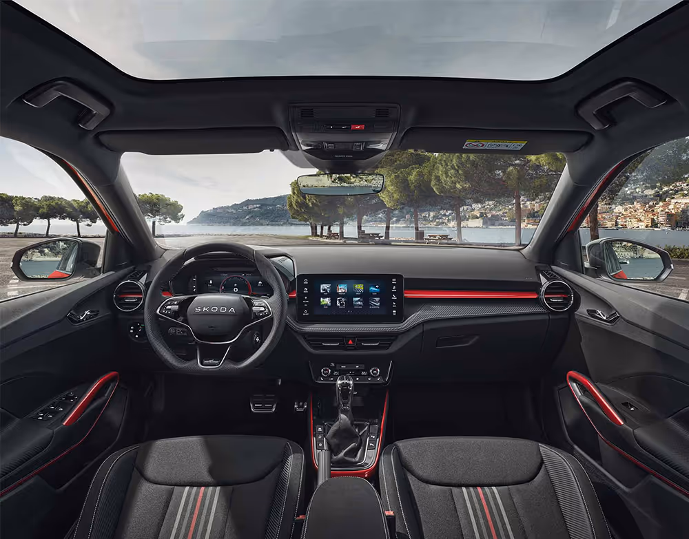 Eine weite Innenansicht zeigt das gesamte Cockpit eines Škoda Fabia mit der Design Selection Monte Carlo. Das dynamische Interieur besticht durch Sportsitze in Schwarz-Rot mit Carbon-Optik-Dekor und markanten roten Akzentleisten am Armaturenbrett. Ein Sportlederlenkrad mit roten Nähten und der Infotainmentbildschirm runden das Design ab. Durch die Windschutzscheibe ist eine malerische Küstenlandschaft mit Bäumen und einem Gewässer zu sehen.