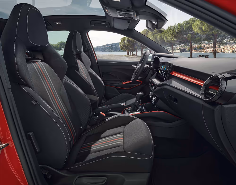 Eine seitliche Innenansicht des Škoda Fabia zeigt das Interieur der Design Selection Monte Carlo. Die Sportsitze in Schwarz mit roten Applikationen und Carbon-Optik sowie das Armaturenbrett mit einer markanten roten Dekorleiste sind zu sehen, während durch die Fenster eine helle Küstenlandschaft erkennbar ist.