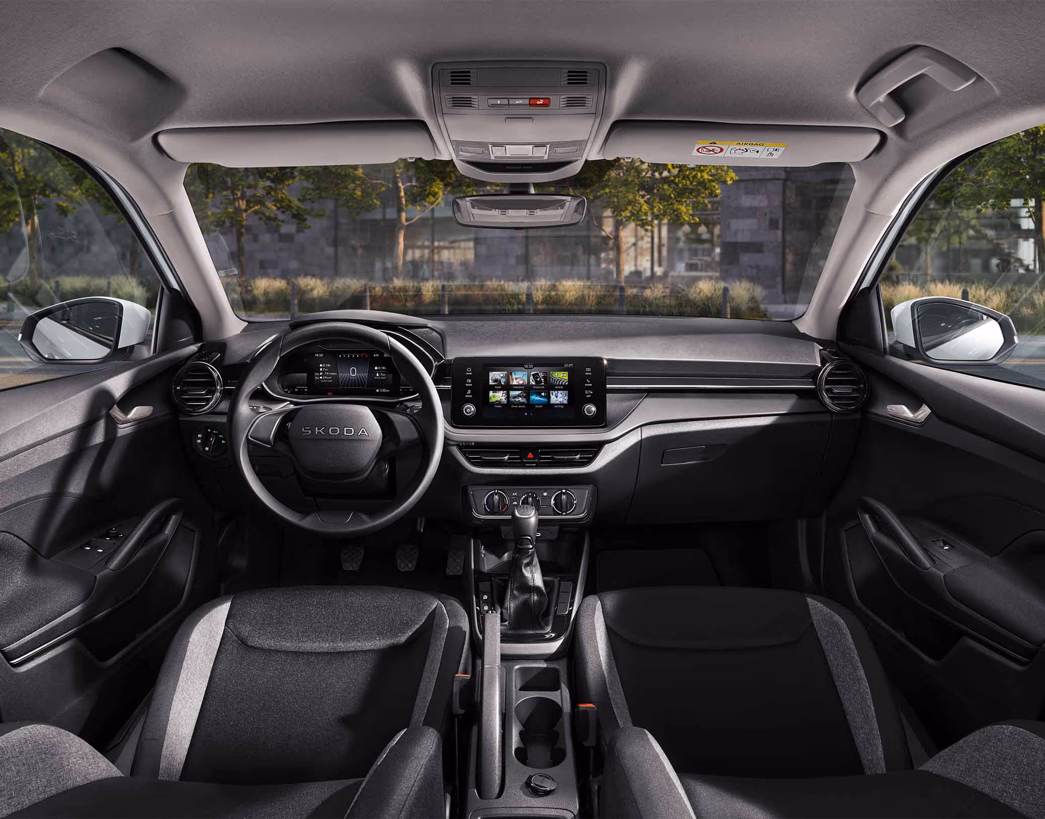 Eine weite Innenansicht zeigt das gesamte Cockpit eines Škoda Fabia mit der Design Selection Studio. Das puristische Interieur verfügt über schwarze Stoffsitze mit grauem Muster und Standard-Dekorelemente in Schwarz. Ein Infotainmentbildschirm und das Lenkrad stehen im Fokus des aufgeräumten Armaturenbretts. Durch die Windschutzscheibe ist eine städtische Kulisse mit Bäumen und einer Eisenbahnbrücke aus Naturstein zu sehen.