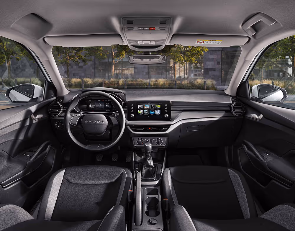 Eine weite Innenansicht zeigt das gesamte Cockpit eines Škoda Fabia mit der Design Selection Studio. Das puristische Interieur verfügt über schwarze Stoffsitze mit grauem Muster und Standard-Dekorelemente in Schwarz. Ein Infotainmentbildschirm und das Lenkrad stehen im Fokus des aufgeräumten Armaturenbretts. Durch die Windschutzscheibe ist eine städtische Kulisse mit Bäumen und einer Eisenbahnbrücke aus Naturstein zu sehen.