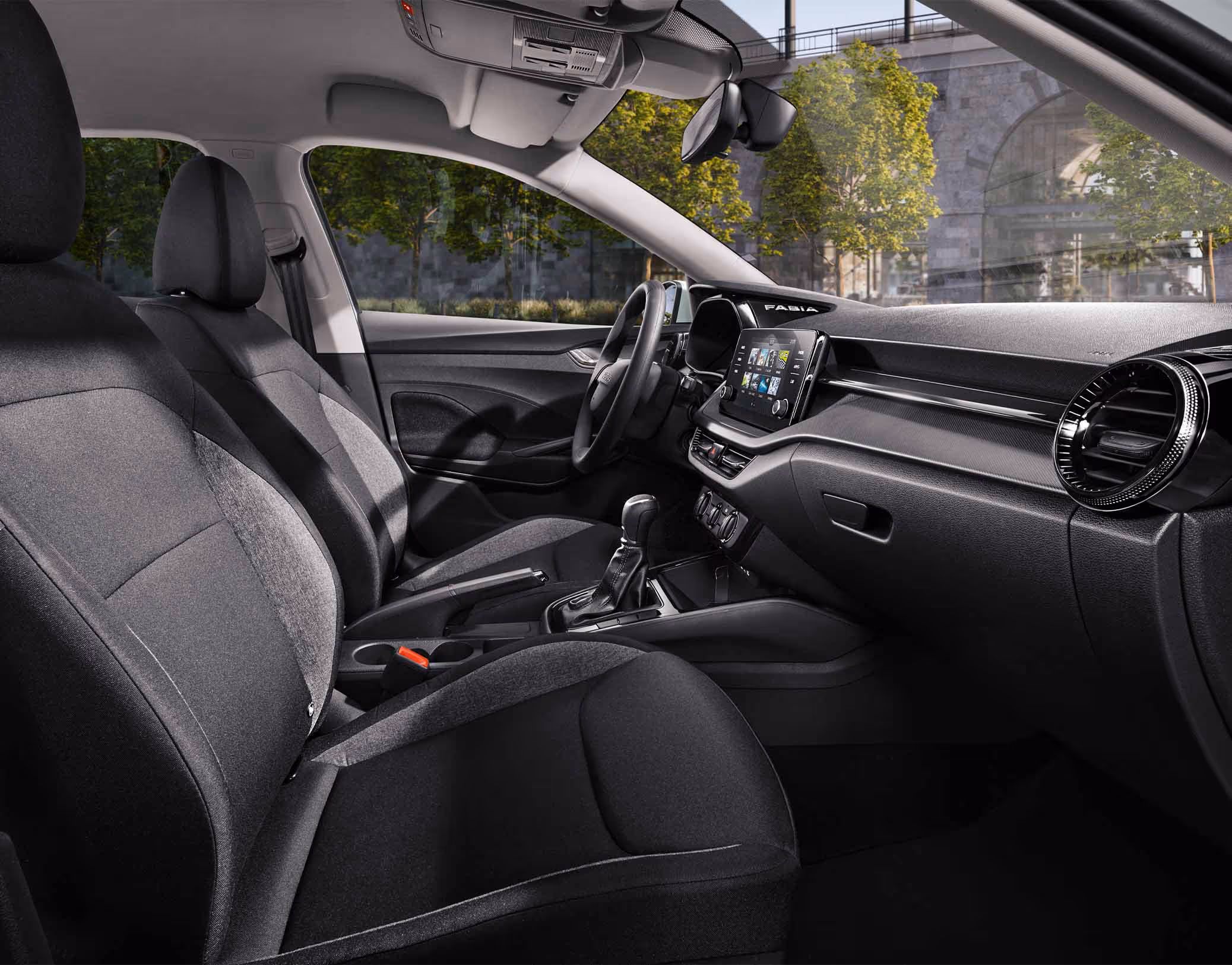 Eine seitliche Innenansicht des Škoda Fabia zeigt das Interieur der Design Selection Studio. Die schwarzen Stoffsitze mit dezentem grauem Muster sowie das Armaturenbrett mit schwarzen Standard-Dekorelementen sind zu sehen, während durch die Fenster eine städtische Kulisse mit Bäumen und einer Eisenbahnbrücke aus Naturstein sichtbar ist.