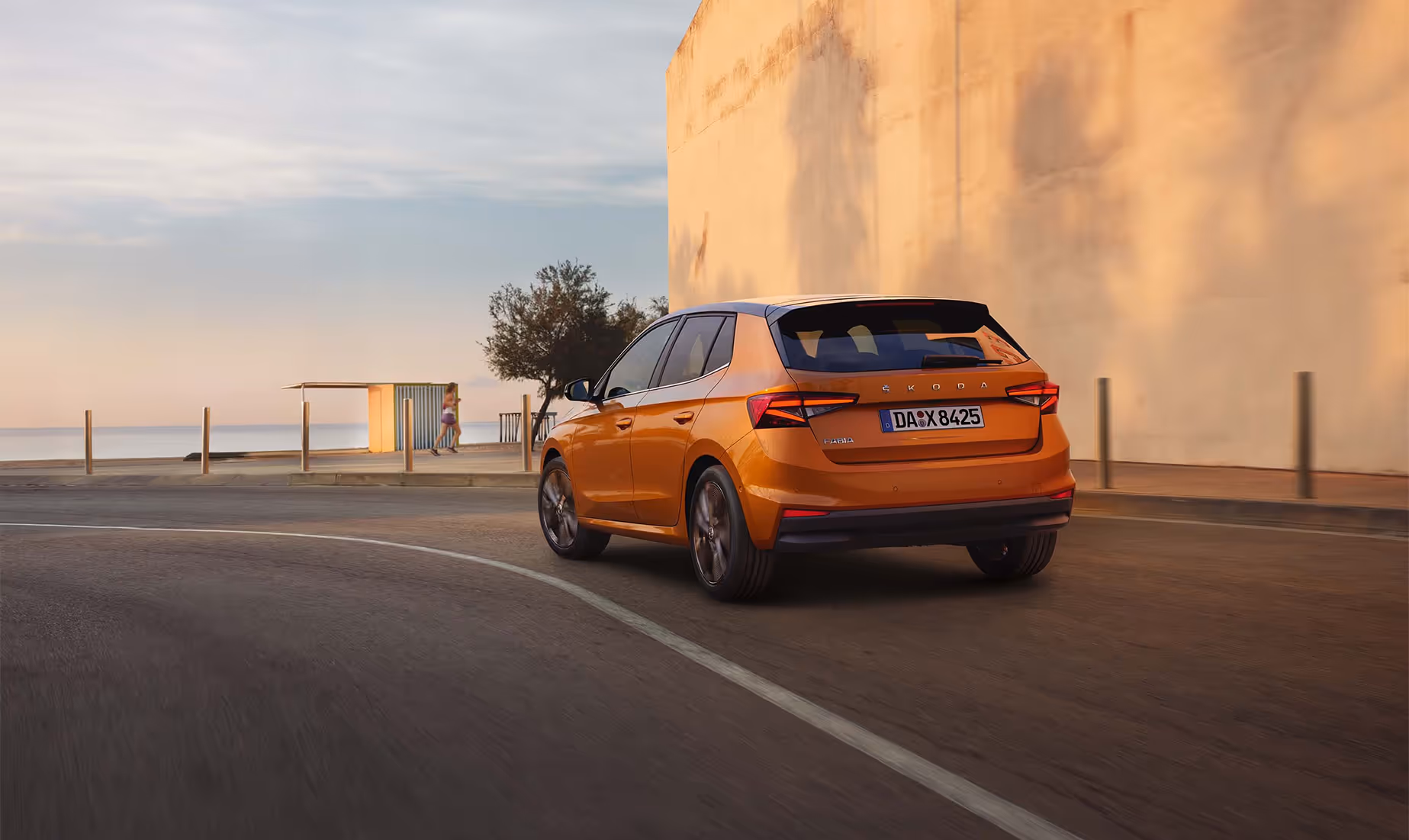 Heckansicht eines Škoda Fabia in der Lackierung Phoenix-Orange Metallic, der dynamisch auf einer Küstenstraße fährt. Das Fahrzeug ist mit dem Color Concept ausgestattet, erkennbar am schwarz lackierten Dach und den schwarzen Außenspiegelkappen. Im Hintergrund sind bei weichem Abendlicht das Meer, ein kleiner Strandpavillon und eine spazierende Person zu sehen, während auf der rechten Seite eine hohe, helle Mauer die Straße säumt.