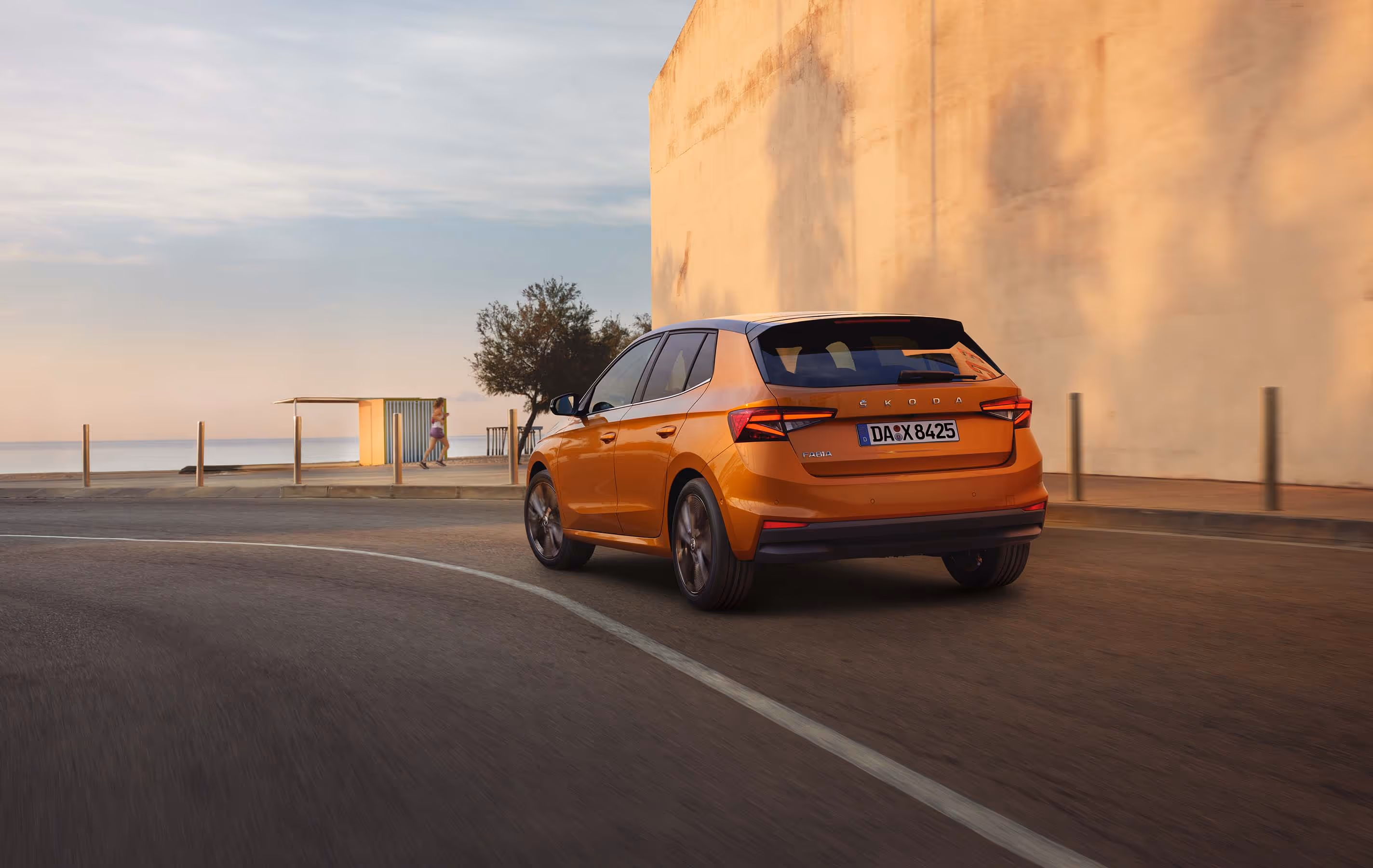 Heckansicht eines Škoda Fabia in der Lackierung Phoenix-Orange Metallic, der dynamisch auf einer Küstenstraße fährt. Das Fahrzeug ist mit dem Color Concept ausgestattet, erkennbar am schwarz lackierten Dach und den schwarzen Außenspiegelkappen. Im Hintergrund sind bei weichem Abendlicht das Meer, ein kleiner Strandpavillon und eine spazierende Person zu sehen, während auf der rechten Seite eine hohe, helle Mauer die Straße säumt.