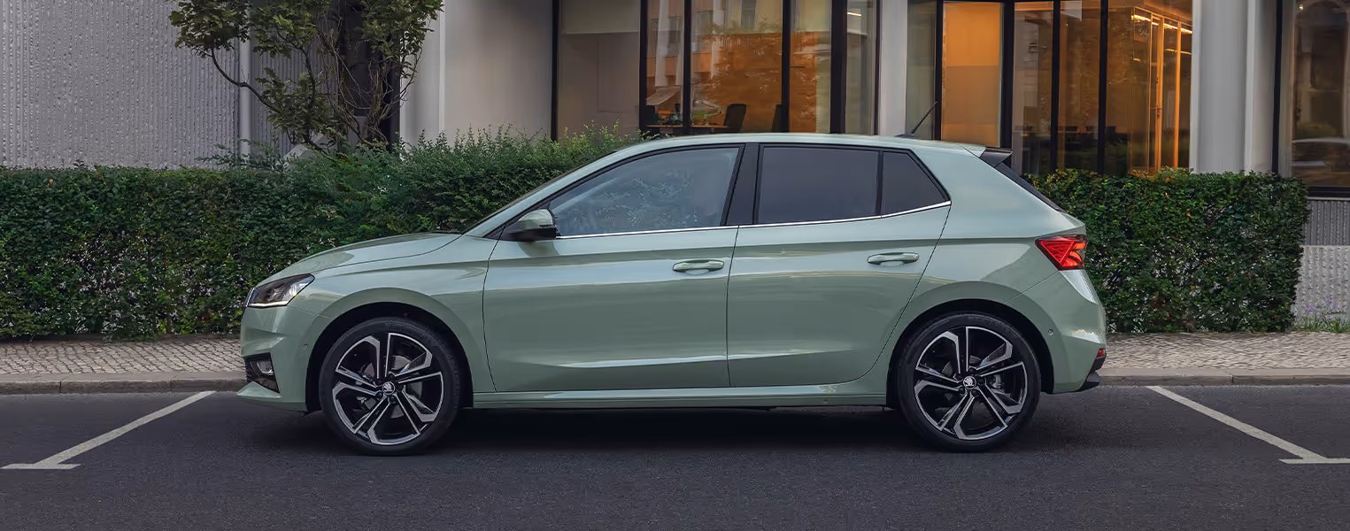 Eine Seitenansicht des Škoda Fabia in der Lackierung Timiano-Grün hebt die dynamische Linienführung und die ausgewogenen Proportionen des Fahrzeugs hervor. Der Wagen steht geparkt vor einer grünen Hecke und einem modernen Gebäude mit beleuchteten Fenstern. Die markanten Leichtmetallfelgen unterstreichen dabei den sportlich-eleganten Auftritt des Kleinwagens.