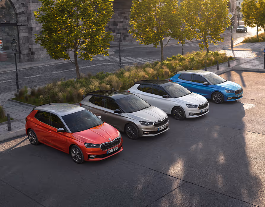 Eine Aufnahme aus der Vogelperspektive zeigt vier Škoda Fabia Modelle, die diagonal versetzt auf einem städtischen Platz stehen. Die Fahrzeugflotte präsentiert verschiedene Lackierungen wie Velvet-Rot Metallic, Graphite-Grau Metallic, Moon-Weiß Perleffekt und Race-Blau Metallic, die jeweils mit einem kontrastierenden Dach in Schwarz oder Grau (Colour Concept) kombiniert sind. Das warme Licht der tiefstehenden Sonne wirft lange Schatten auf das Kopfsteinpflaster und die umgebenden Bäume.