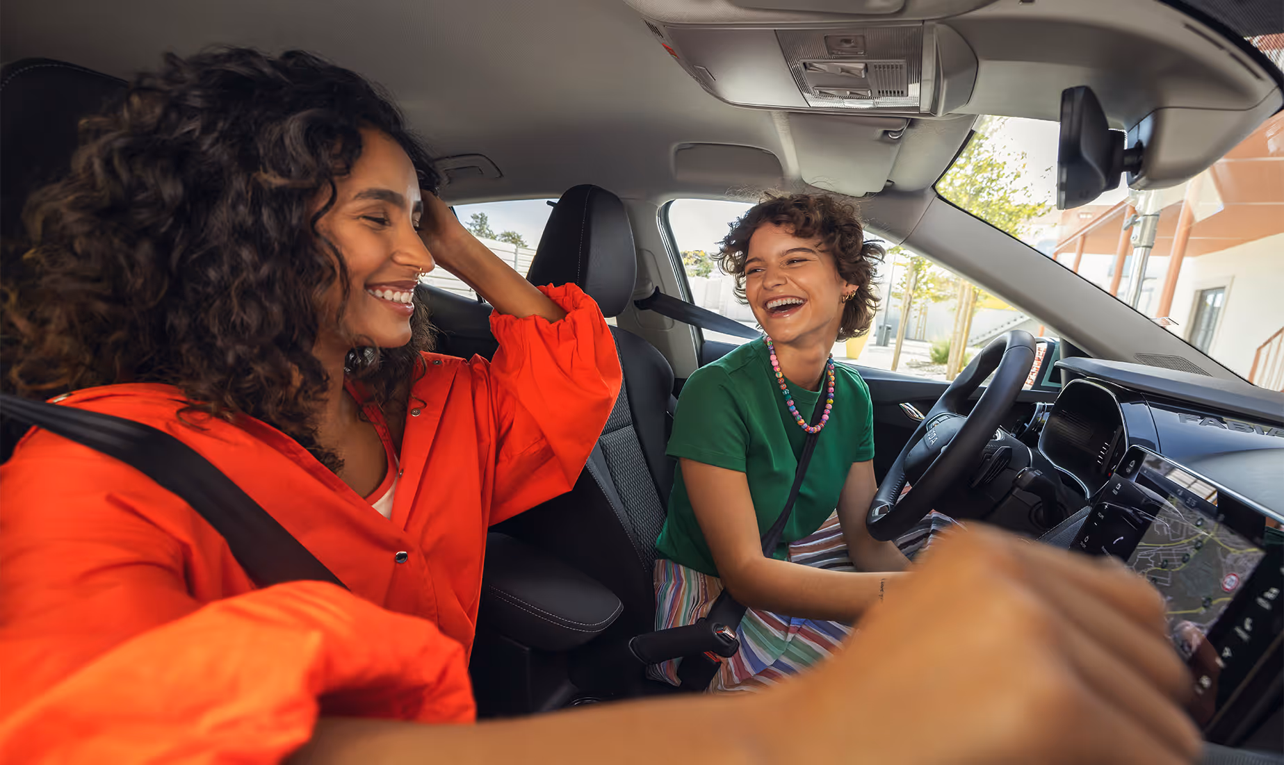 Zwei junge Frauen sitzen lachend im Interieur des Škoda Fabia und strahlen ausgelassene Lebensfreude aus. Der Fokus liegt auf dem modernen Cockpit mit dem griffigen Lenkrad und dem zentralen Infotainment-Display, auf dem die Navigation zu sehen ist. Die fröhliche Atmosphäre unterstreicht das angenehme Raumgefühl und die Konnektivität des Fahrzeugs.
