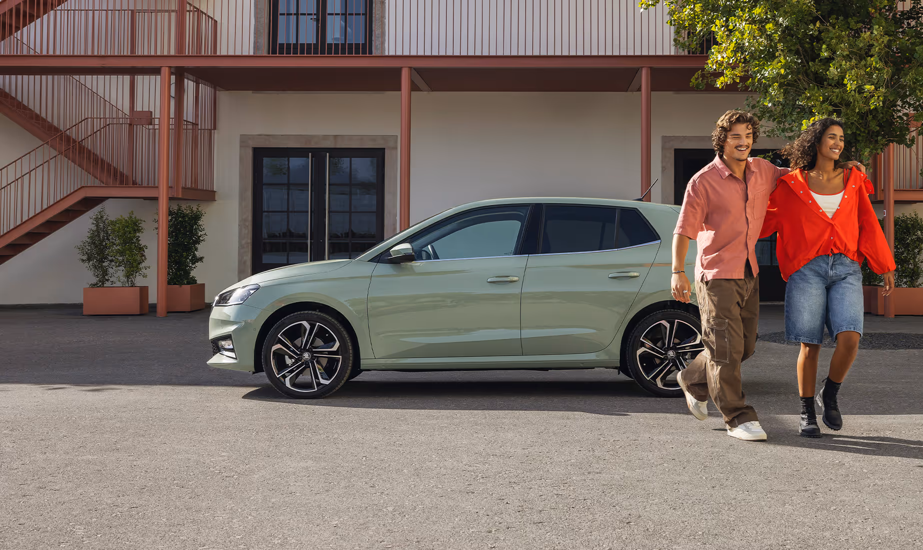 Ein Škoda Fabia in der Lackierung Timiano-Grün steht geparkt vor einem Gebäude mit markanter, terrakottafarbener Außenstruktur. Ein junges Paar spaziert lachend und Arm in Arm am Fahrzeug vorbei, was die urbane Lebensfreude unterstreicht. Die Seitenansicht hebt die elegante Silhouette des Kleinwagens sowie die dynamischen Leichtmetallfelgen hervor.