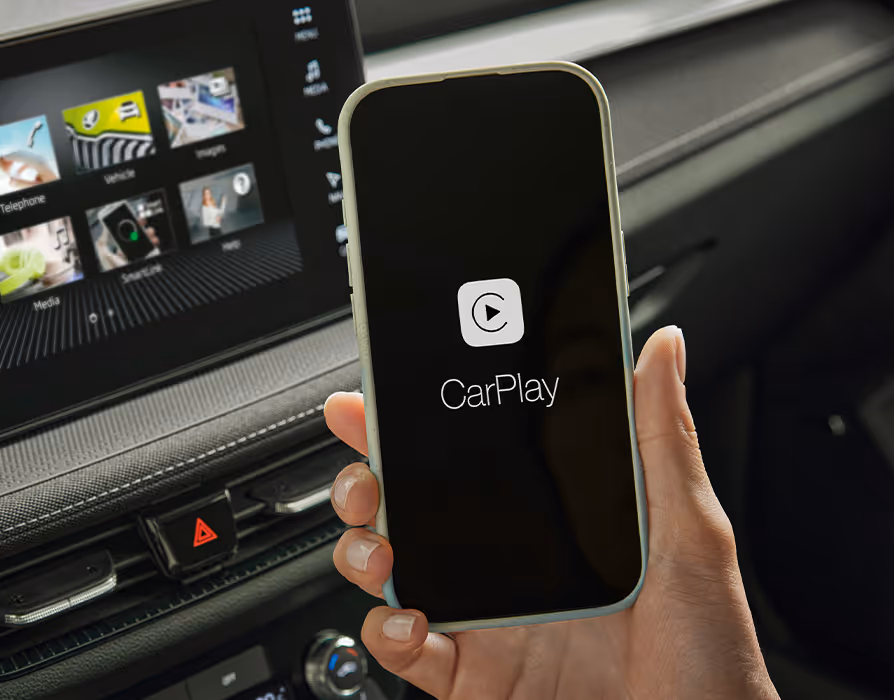 Die Nahaufnahme präsentiert eine Hand, die ein Smartphone mit dem CarPlay Logo vor dem Armaturenbrett hält. Im Hintergrund ist das aktivierte Infotainment-Display mit Menüansicht sowie die darunter liegende Leiste mit Lüftungsdüsen und Warnblinkschalter zu sehen.