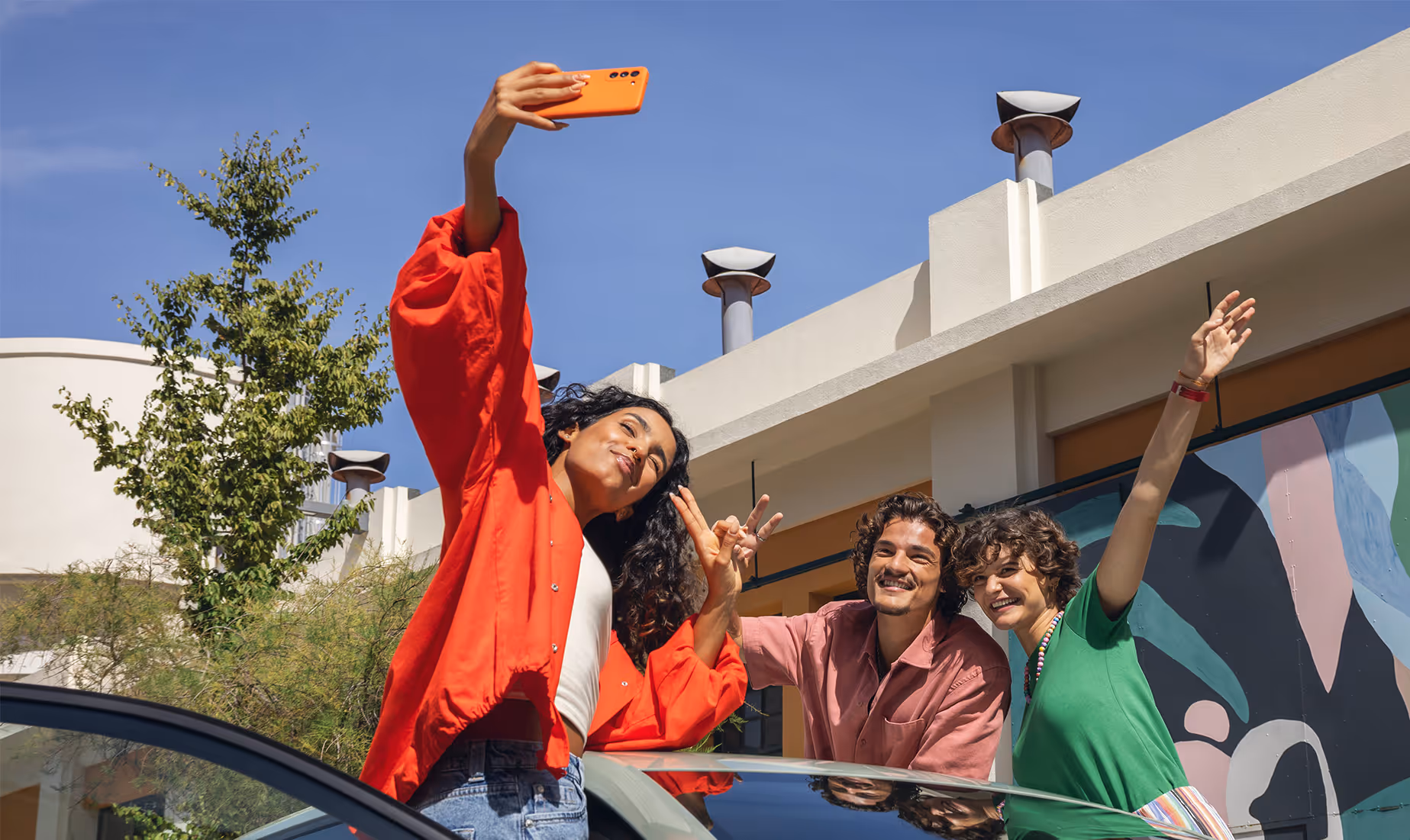 Drei junge Erwachsene machen bei strahlendem Sonnenschein ein ausgelassenes Selfie, während sie am Škoda Fabia in der Lackierung Timiano-Grün stehen. Von dem Fahrzeug ist die Dachlinie zu erkennen, die den unteren Bildrand bildet. Die Szene vor einem Gebäude mit blauem Himmel im Hintergrund vermittelt pure Lebensfreude und einen vernetzten, modernen Lifestyle.