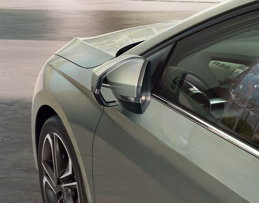 Eine Detailaufnahme von schräg oben zeigt den angeklappten Außenspiegel auf der Fahrerseite des Škoda Fabia in der Lackierung Timiano-Grün. Das Spiegelgehäuse in Wagenfarbe ist mit einem schmalen, integrierten Blinkerstreifen ausgestattet. Durch die Seitenscheibe fällt der Blick auf den seitlichen „FABIA“-Schriftzug der Instrumententafel sowie einen Teil des digitalen Cockpits.