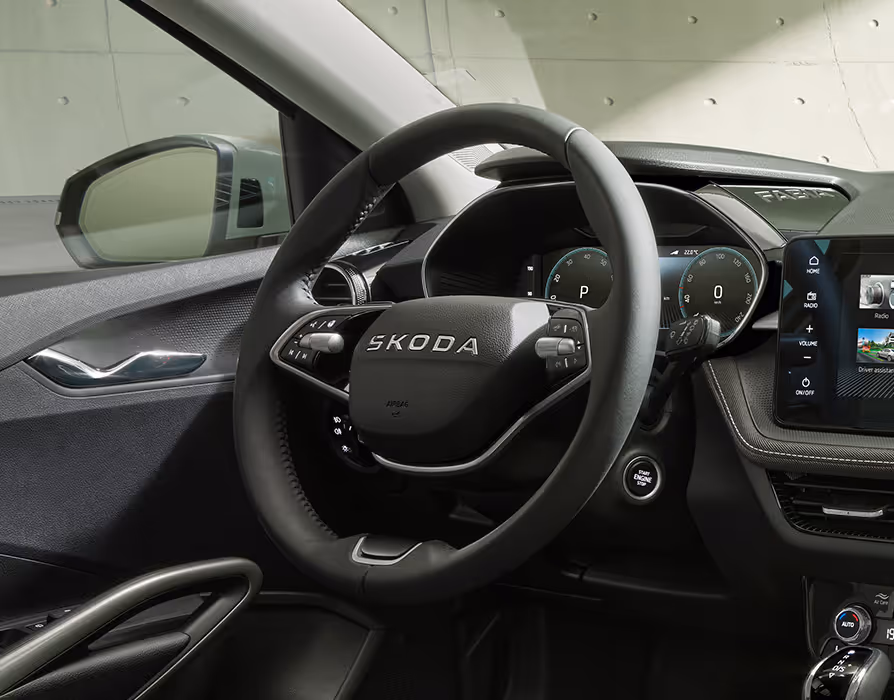Eine Nahaufnahme des Fahrerplatzes rückt das Multifunktionslederlenkrad mit seinen verchromten Bedienelementen und dem zentralen ŠKODA-Schriftzug in den Mittelpunkt. Dahinter ist das digitale Kombiinstrument (Virtual Cockpit) ablesbar, während rechts der Infotainment-Touchscreen und der Start-Stopp-Knopf an der Lenksäule zu sehen sind. Linkerhand sind Teile der Türverkleidung mit dem verchromten Türöffner sowie der Blick in den Außenspiegel erkennbar.