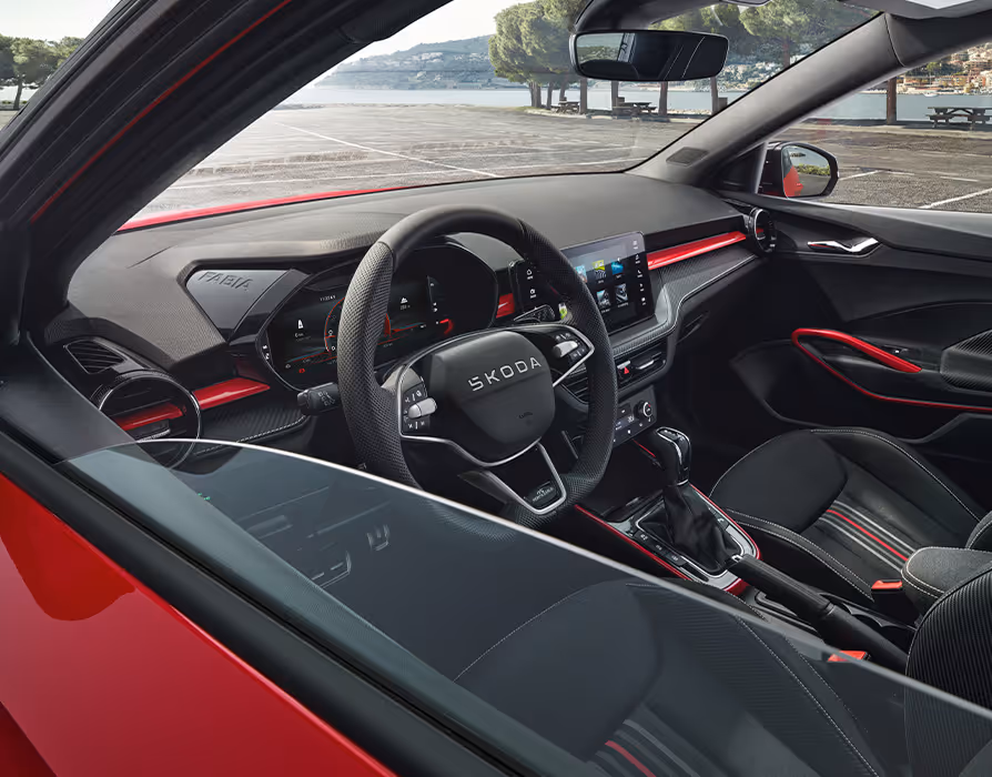 Ein Blick in den Innenraum des Škoda Fabia Monte Carlo zeigt das sportliche Cockpit mit markanten roten Dekorleisten an Armaturenbrett, Mittelkonsole und Türgriffen. Im Fokus steht das perforierte Multifunktions-Sportlenkrad mit abgeflachtem Kranz vor dem digitalen Kombiinstrument. Die schwarzen Sportsitze mit hellem Streifendesign und integrierten Kopfstützen komplettieren die Ansicht.