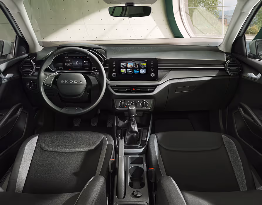 Ein Blick in das Interieur des Škoda Fabia der Ausstattungsvariante Essence präsentiert die Design Selection Studio mit ihren charakteristischen Stoffbezügen in Schwarz und Grau. Das Cockpit umfasst ein schlichtes zweispeichiges Lenkrad, ein zentrales Infotainment-Display sowie die Bedienelemente der manuellen Klimaanlage. Die Mittelkonsole ist mit einem manuellen Schalthebel, einer mechanischen Handbremse und offenen Getränkehaltern ausgestattet.