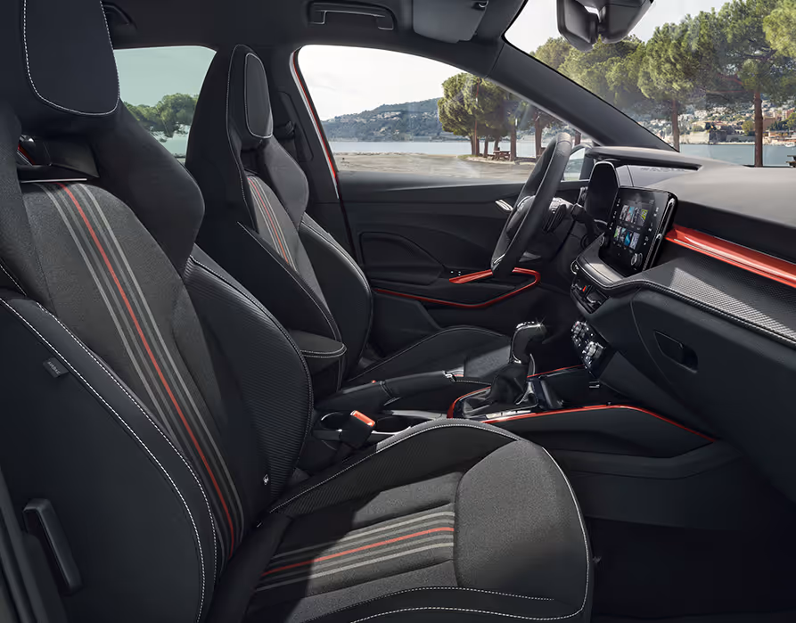 Eine Seitenansicht der Vordersitze zeigt die schwarzen Sportsitze mit integrierten Kopfstützen und markantem Streifendesign im exklusiven Interieur der Design Selection Monte Carlo. Rote Dekorleisten an Armaturenbrett, Mittelkonsole und Türgriffen setzen starke Farbakzente im Kontrast zu Elementen in Carbon-Optik. Das abgeflachte Sportlenkrad und das digitale Cockpit vervollständigen die sportliche Ausstattung.