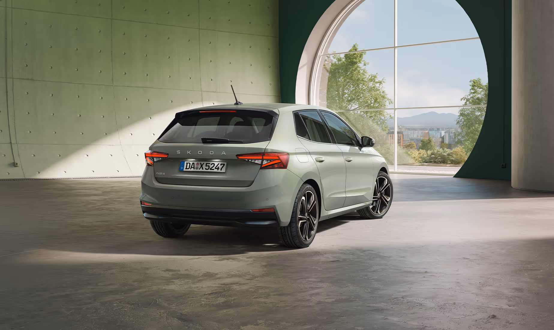 Das Heck des Škoda Fabia in der Lackierung Timiano-Grün präsentiert sich in einem modernen Raum mit Sichtbetonwänden und einem großen runden Fenster. Das Tageslicht setzt die markanten Rückleuchten, den mittigen „ŠKODA“-Schriftzug auf der Heckklappe sowie die sportlichen Leichtmetallfelgen in Szene. Der Blickwinkel von schräg hinten unterstreicht die dynamische Linienführung und das kompakte Design des Fahrzeugs.