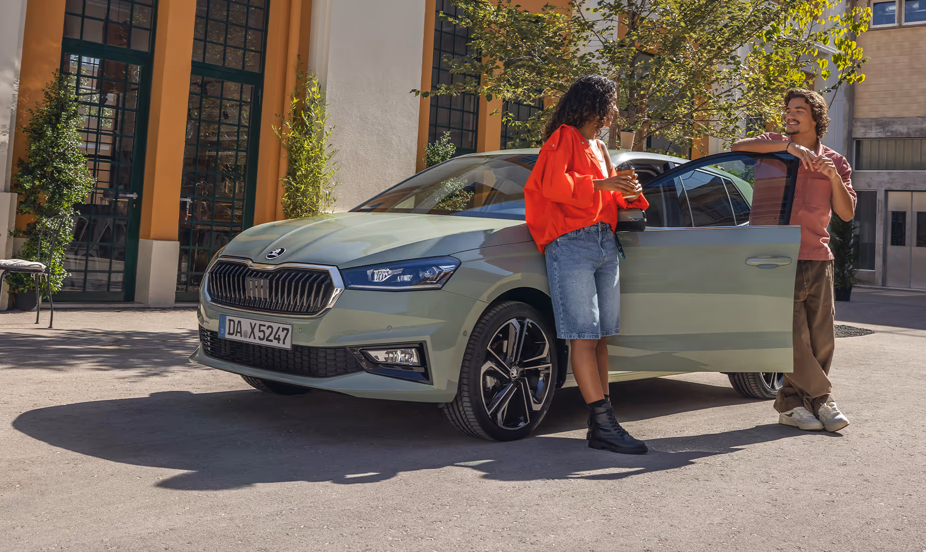 Ein Škoda Fabia in der Lackierung Timiano-Grün steht in einer sonnigen, städtischen Kulisse vor einem Gebäude mit großen Fenstertüren. Eine Frau lehnt entspannt mit einem Kaffeebecher am vorderen Kotflügel, während ein Mann an der geöffneten Fahrertür steht und sich mit ihr unterhält. Die Szene setzt das moderne Design des Kleinwagens und die markanten Leichtmetallfelgen in einen lebendigen Alltagskontext.