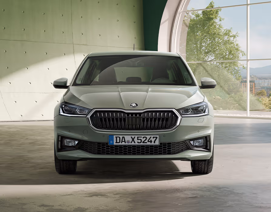 Der Škoda Fabia in der Lackierung Timiano-Grün präsentiert sich in der direkten Frontalansicht in einem modernen Raum mit Sichtbetonwänden. Der markante Kühlergrill mit Chromrahmen und die scharf gezeichneten Scheinwerfer prägen das selbstbewusste Gesicht des Kleinwagens. Das durch das große Rundfenster seitlich einfallende Licht betont die Linienführung der Motorhaube und der Frontschürze.