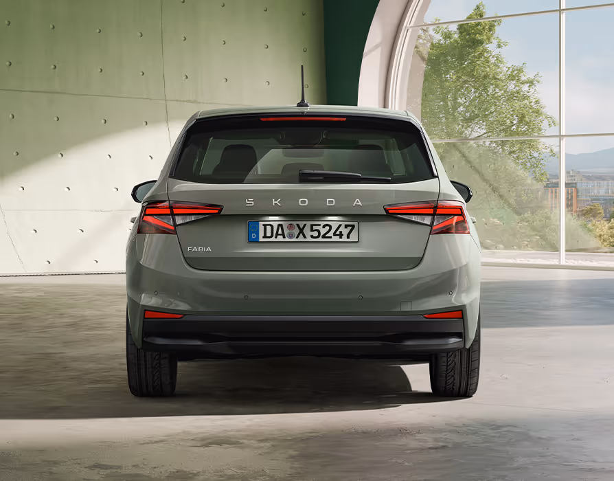 Der Škoda Fabia in der Lackierung Timiano-Grün ist in der direkten Heckansicht vor einer modernen Sichtbetonwand zu sehen. Im Mittelpunkt stehen der prominente „ŠKODA“-Schriftzug auf der Heckklappe und die kristallinen Rückleuchten. Die klare horizontale Gliederung und der dunkle Diffusorbereich am unteren Stoßfänger unterstreichen den breiten Stand des Fahrzeugs.