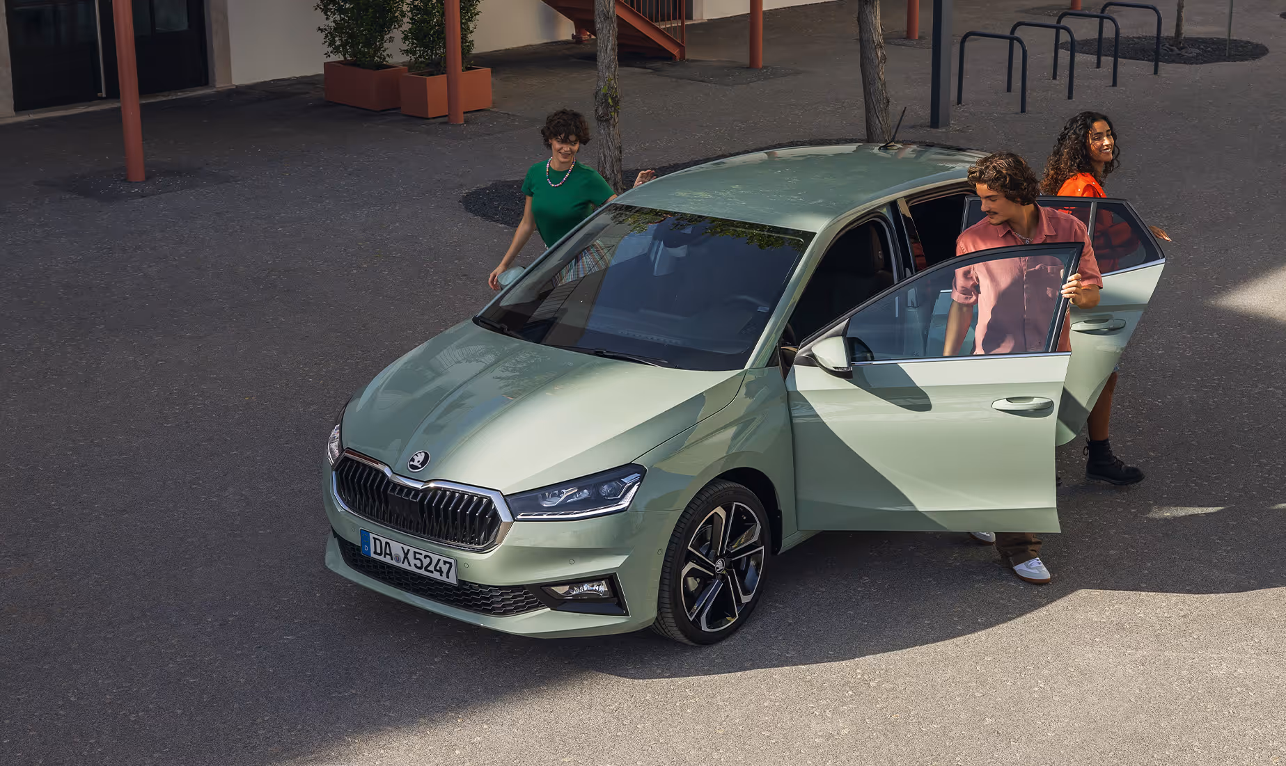 Aus der Vogelperspektive ist ein Škoda Fabia in der Lackierung Timiano-Grün zu sehen, aus dem gerade drei junge Erwachsene aussteigen. Die Szene auf einem sonnigen Platz unterstreicht die Geräumigkeit und die soziale Komponente des Kleinwagens. Das Lichtspiel auf der Karosserie hebt dabei die elegante Form und die sportlichen Leichtmetallfelgen hervor.