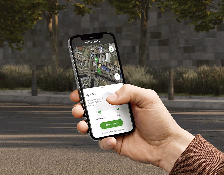 Eine Nahaufnahme zeigt eine Hand, die ein Smartphone mit der geöffneten My ŠKODA App hält. Auf dem Display wird der aktuelle Standort des „My FABIA“ über die Funktion „Parking Position“ auf einer Karte dargestellt. Der Hintergrund lässt eine unscharfe städtische Umgebung mit Bäumen und einem Gebäude erkennen.