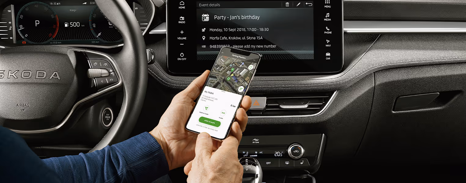 Eine Ansicht aus der Fahrerperspektive zeigt ein Smartphone mit der geöffneten My ŠKODA App, auf der die Parkposition des Fahrzeugs dargestellt wird. Im Hintergrund sind das digitale Kombiinstrument (Virtual Cockpit) hinter dem Lenkrad sowie das zentrale Infotainment-Display mit angezeigten Kalenderdetails zu sehen. Darunter befinden sich die Bedienelemente der Klimaanlage und der Wählhebel des Automatikgetriebes.