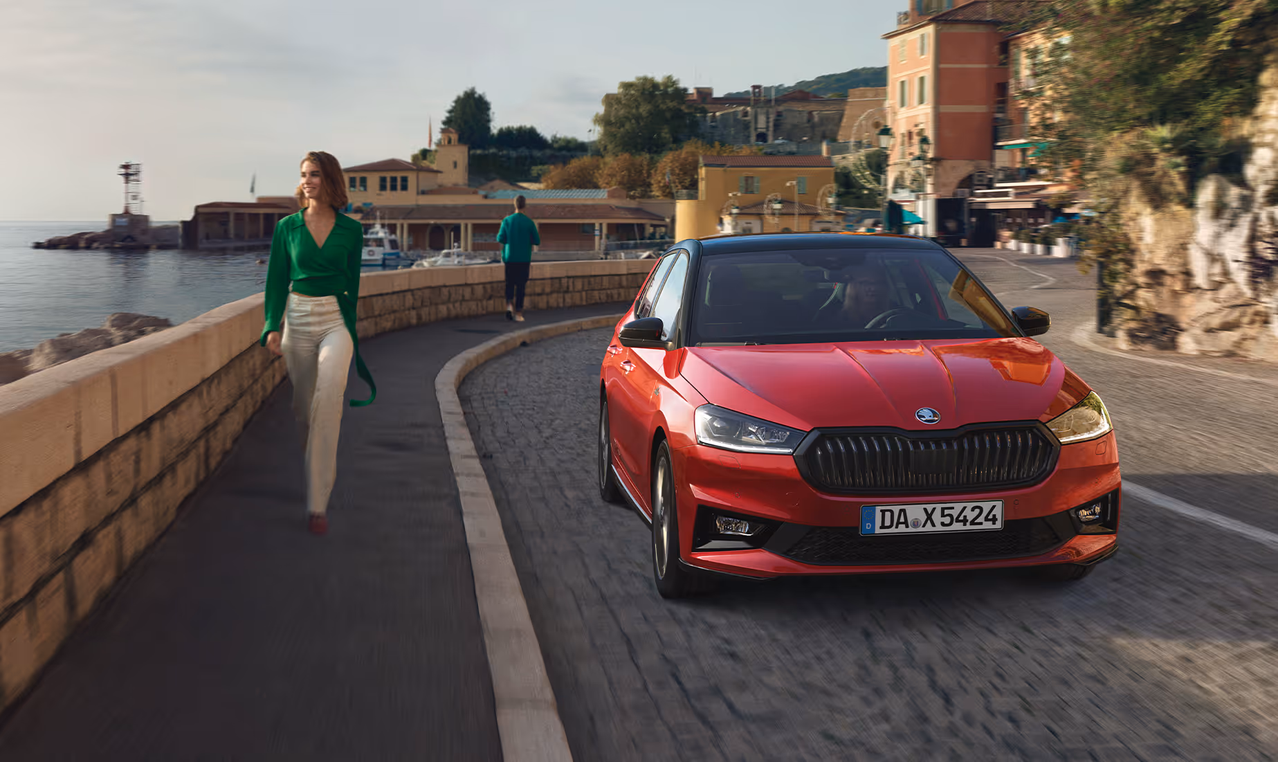 Ein Škoda Fabia Monte Carlo in der Lackierung Velvet-Rot Metallic fährt dynamisch entlang einer sonnigen Küstenstraße mit historischen Gebäuden im Hintergrund. Das Fahrzeug zeichnet sich durch die charakteristischen schwarzen Akzente am Kühlergrill, den Außenspiegeln und dem Dach aus. Auf der angrenzenden Promenade spaziert eine Frau, während links das Meer zu sehen ist.