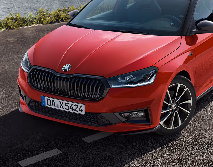 Eine Detailaufnahme der Frontpartie des Škoda Fabia Monte Carlo in Velvet-Rot Metallic zeigt den charakteristischen schwarzen Kühlergrillrahmen und die sportliche Frontschürze mit schwarzen Elementen. Das Bild hebt zudem die scharf gezeichneten LED-Scheinwerfer, die Monte Carlo Plakette am Kotflügel sowie die glanzgedrehten Leichtmetallfelgen hervor. Die dynamische Linienführung der Motorhaube wird durch das einfallende Sonnenlicht zusätzlich betont.