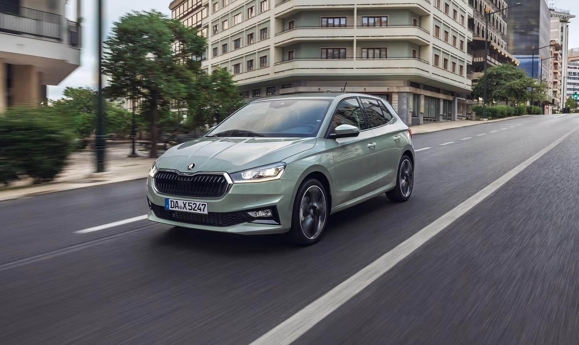 Eine dynamische Fahraufnahme zeigt den Škoda Fabia in der Lackierung Timiano-Grün auf einer mehrspurigen städtischen Straße. Die Bewegung des Fahrzeugs wird durch die Unschärfe der vorbeiziehenden Gebäude und Bäume im Hintergrund sowie der rotierenden Räder verdeutlicht. Die Frontansicht hebt den breiten Kühlergrill und die scharf gezeichneten Scheinwerfer hervor.