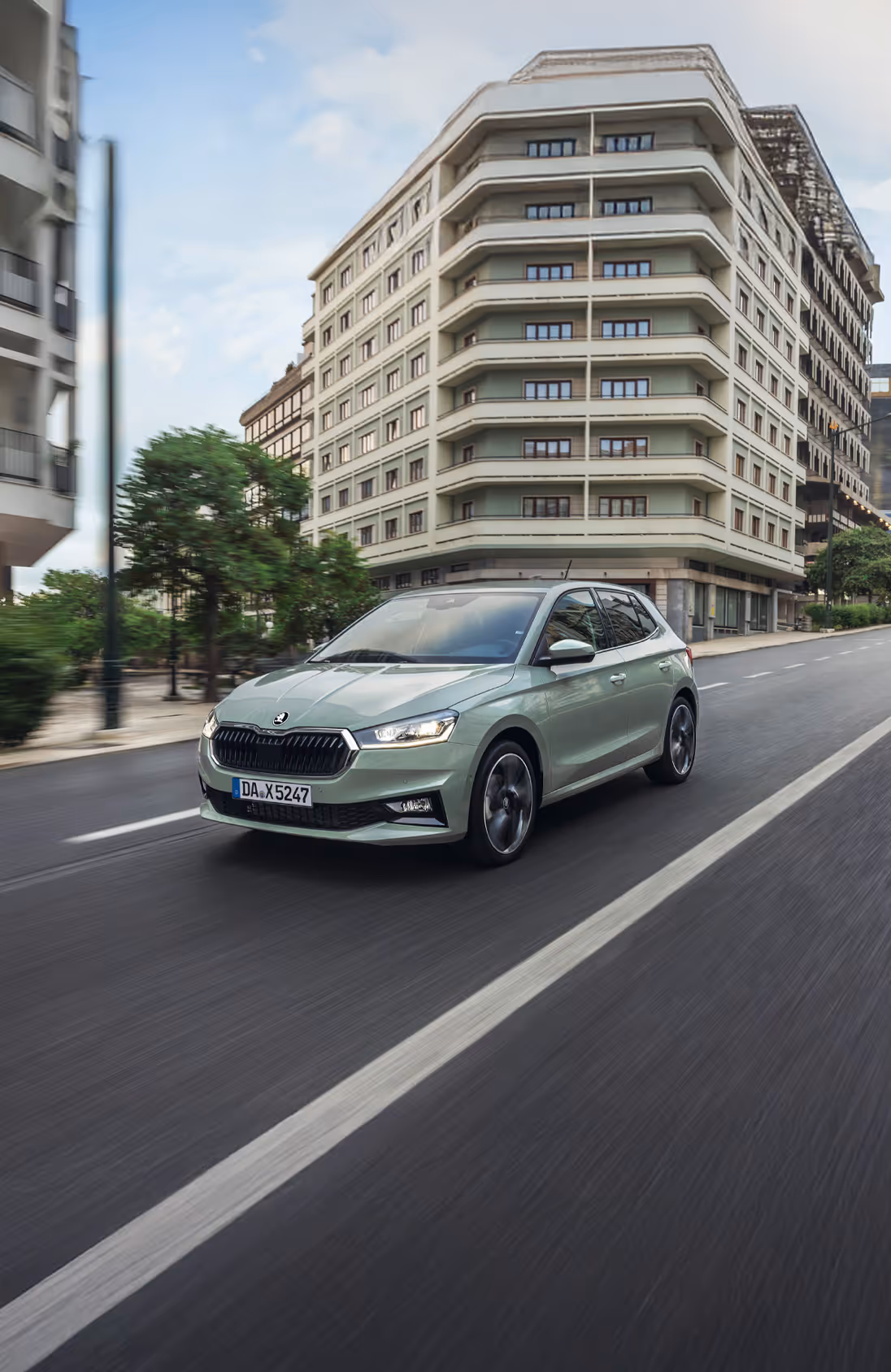 Eine dynamische Fahraufnahme zeigt den Škoda Fabia in der Lackierung Timiano-Grün auf einer mehrspurigen städtischen Straße. Die Bewegung des Fahrzeugs wird durch die Unschärfe der vorbeiziehenden Gebäude und Bäume im Hintergrund sowie der rotierenden Räder verdeutlicht. Die Frontansicht hebt den breiten Kühlergrill und die scharf gezeichneten Scheinwerfer hervor.