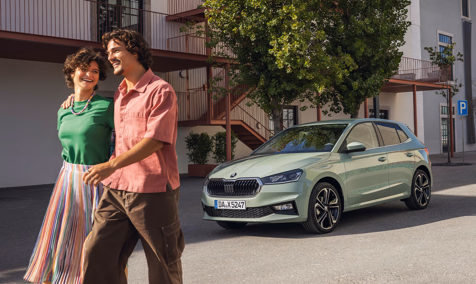 Ein Škoda Fabia in der Lackierung Timiano-Grün steht geparkt vor einem modernen Gebäude mit markanten Außentreppen und begrünten Bereichen. Im Vordergrund spaziert ein junges, lachendes Paar Arm in Arm an dem Fahrzeug vorbei. Die Schrägansicht von vorne präsentiert den Kühlergrill, die Scheinwerfer sowie die dynamische Seitenlinie des Kleinwagens.