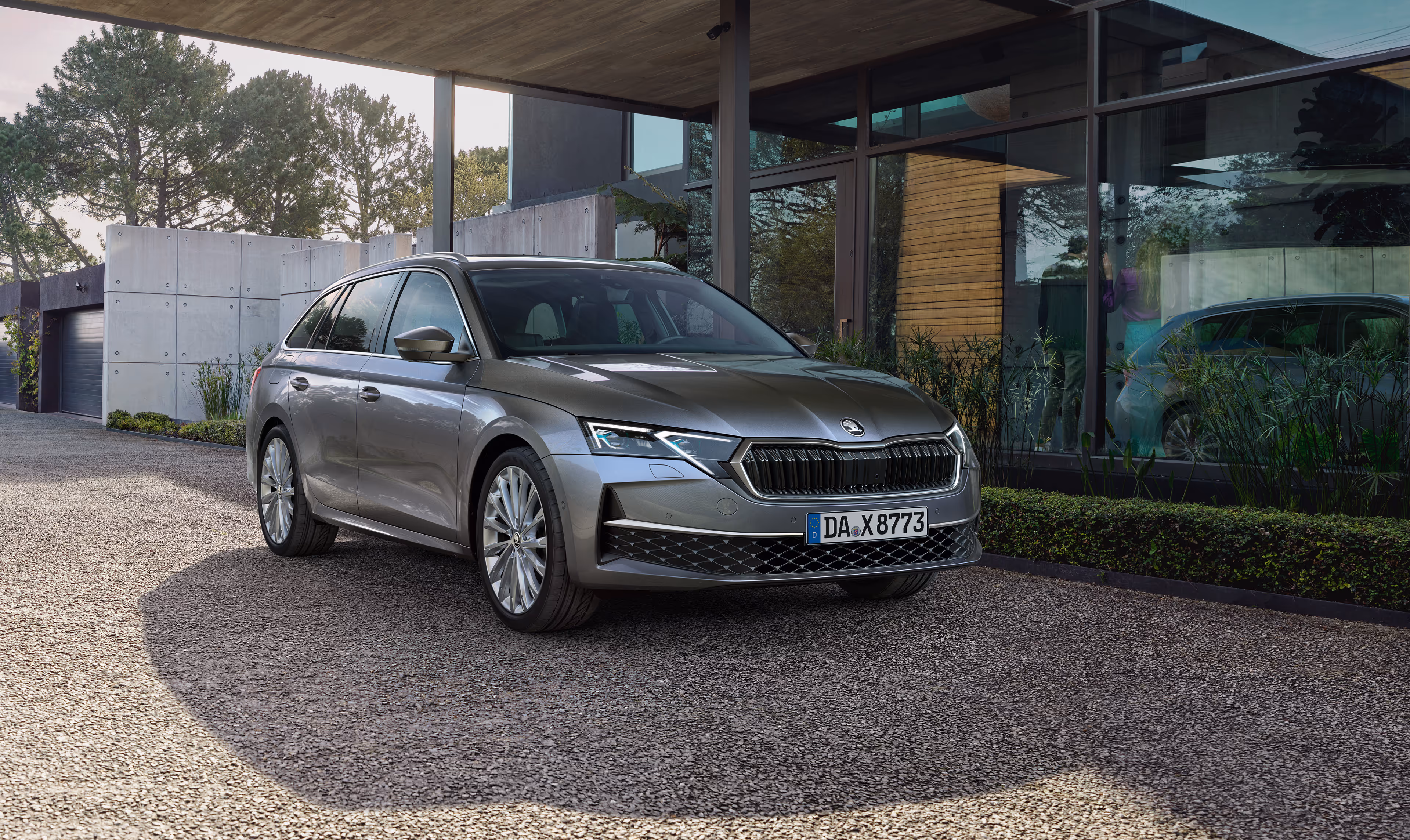 Ein Škoda Octavia Combi in der Lackierung Graphite-Grau Metallic ist in der Kieseinfahrt eines modernen Hauses geparkt. Das Fahrzeug wird in einer Dreiviertelansicht von vorne gezeigt, im Hintergrund sind das Haus mit seiner großen Glasfassade und Bäume zu sehen.