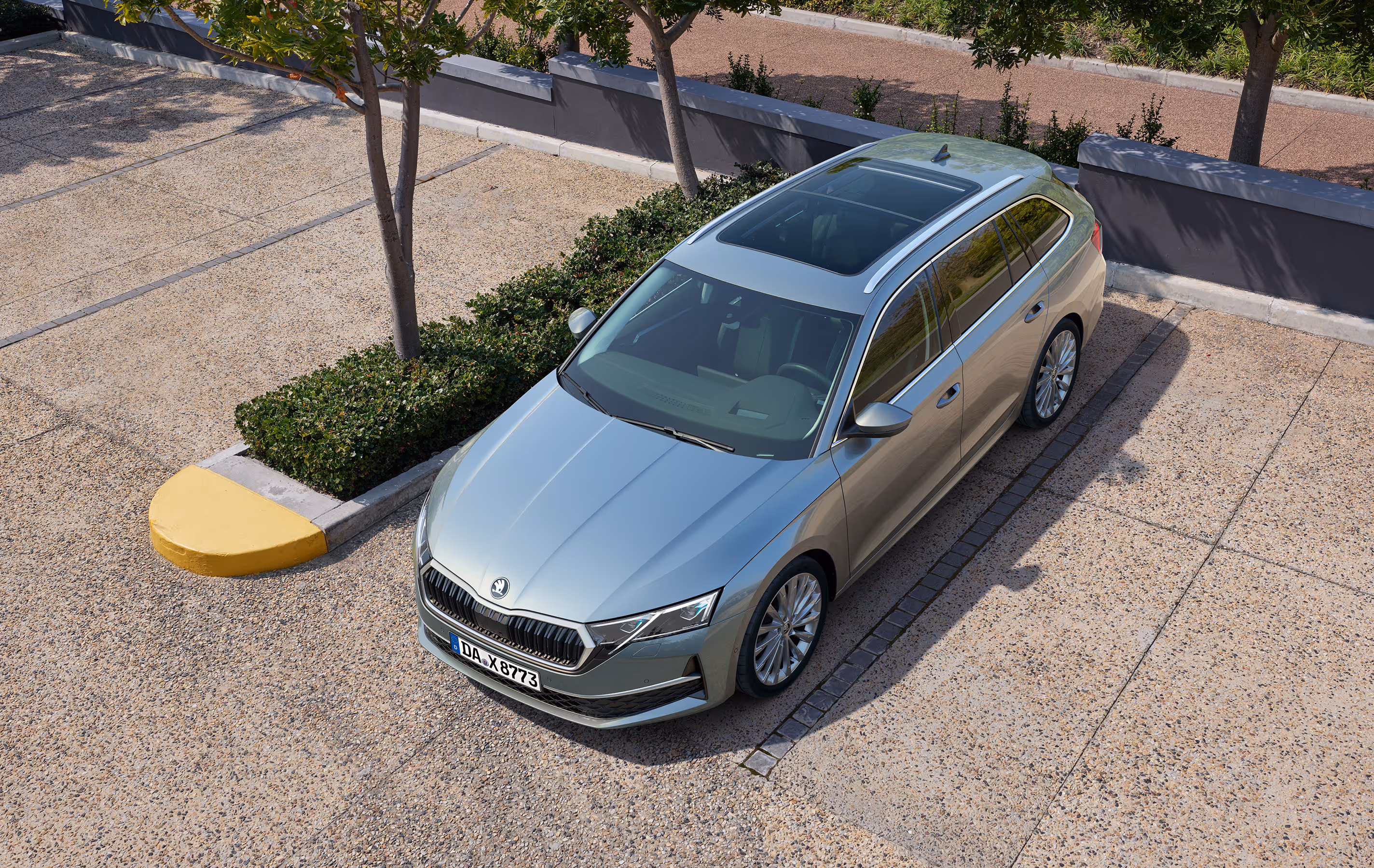 Ein Škoda Octavia Combi in der Lackierung Smokey Diamond-Silber Metallic wird aus einer erhöhten Perspektive auf einem gepflasterten Parkplatz gezeigt. Das Fahrzeug ist mit einem Panoramaschiebedach ausgestattet und parkt neben einem bepflanzten Beet mit jungen Bäumen.