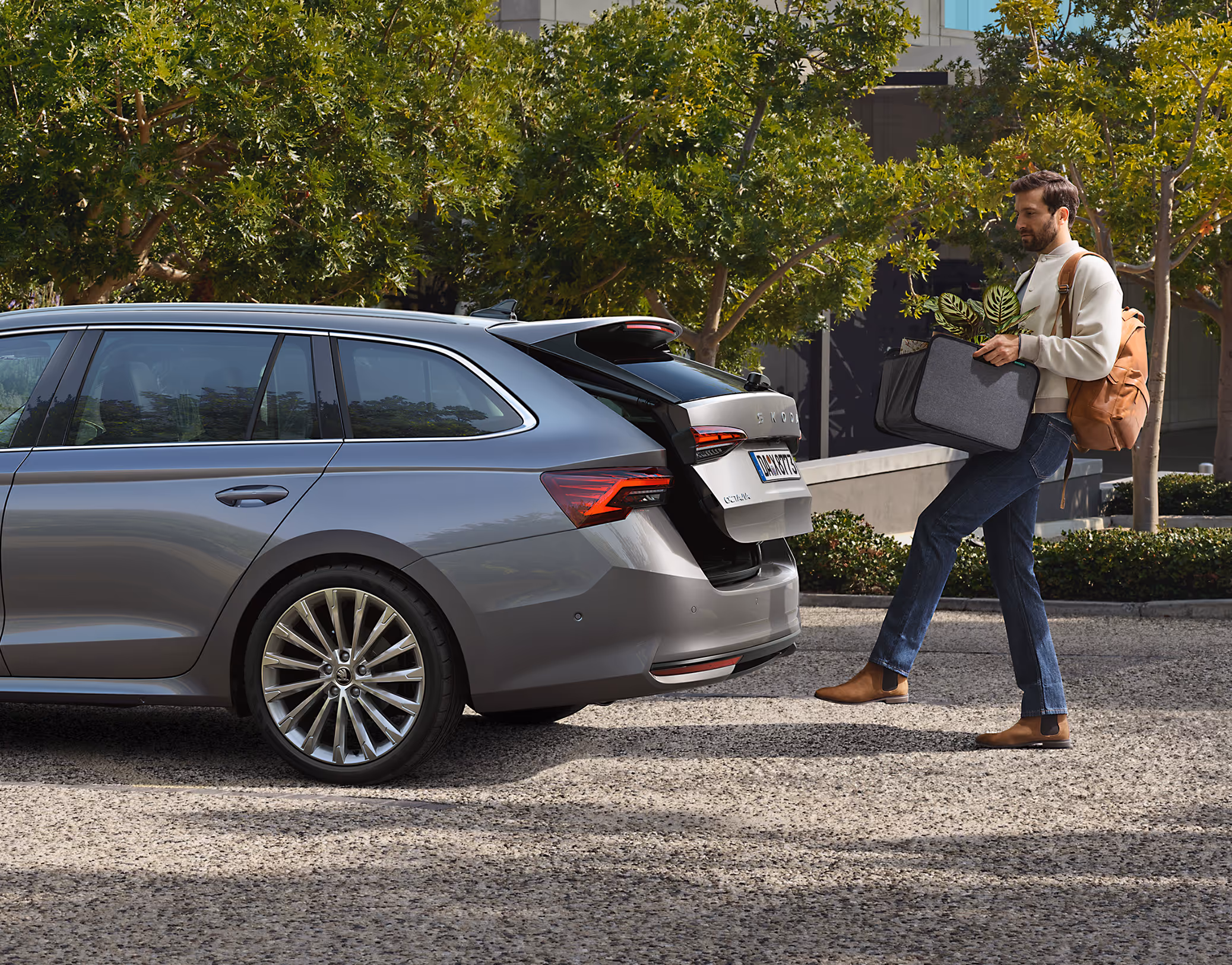 Ein Mann öffnet die elektrische Heckklappe eines Škoda Octavia Combi mit Hilfe des Virtuellen Pedals. Mit einem Rucksack über der Schulter und einer Tasche mit einer Pflanze darin vor sich her tragend löst er mit einer Fußbewegung unter dem Heckstoßfänger den Öffnungsmechanismus aus. Das in Smokey Diamond-Silber Metallic lackierte Fahrzeug ist auf einem gepflasterten Platz geparkt.

