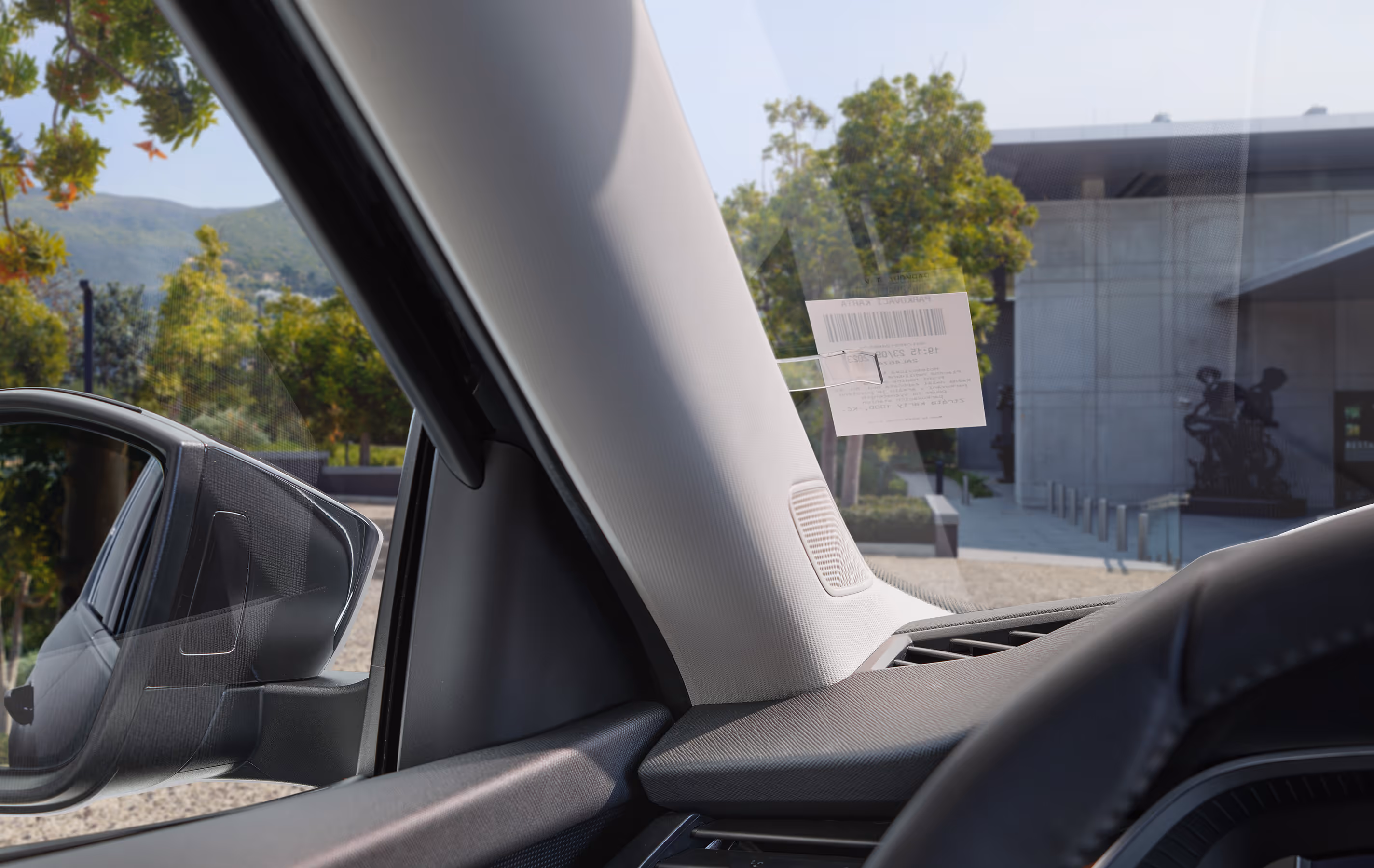 Eine Nahaufnahme aus dem Innenraum eines Škoda Octavia zeigt den Tickethalter an der A-Säule, in dem ein Parkschein steckt. Darunter ist der Lautsprecher des Canton-Soundsystems in die Säulenverkleidung integriert. Durch die Windschutzscheibe ist ein modernes Gebäude mit Bäumen und einer Skulptur davor zu erkennen.