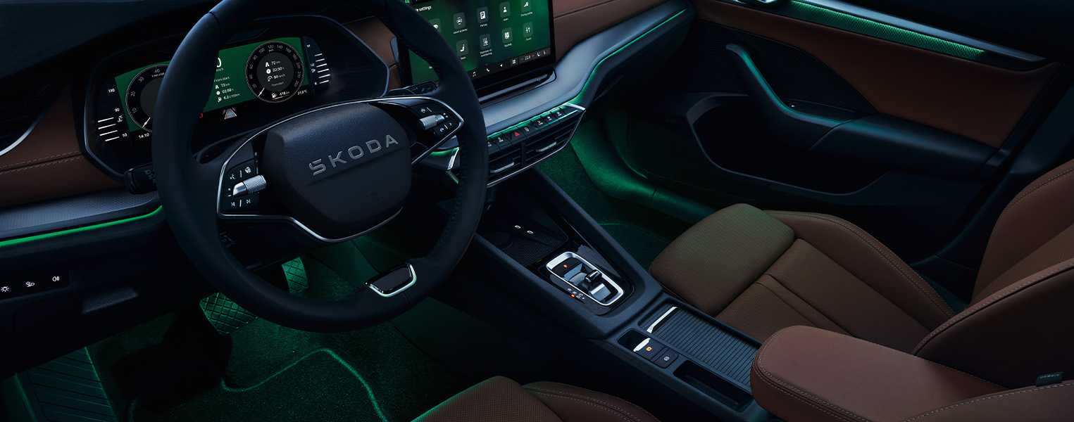 Das Interieur eines Škoda Octavia mit der Design Selection Suite in Cognac wird bei Nacht von der grünen Ambientebeleuchtung in Szene gesetzt. Die Lichtleisten erstrecken sich über das Armaturenbrett und die Türverkleidungen und beleuchten auch den Fußraum. Auf dem digitalen Cockpit und dem Infotainment-Display werden Fahr- und Fahrzeuginformationen angezeigt.